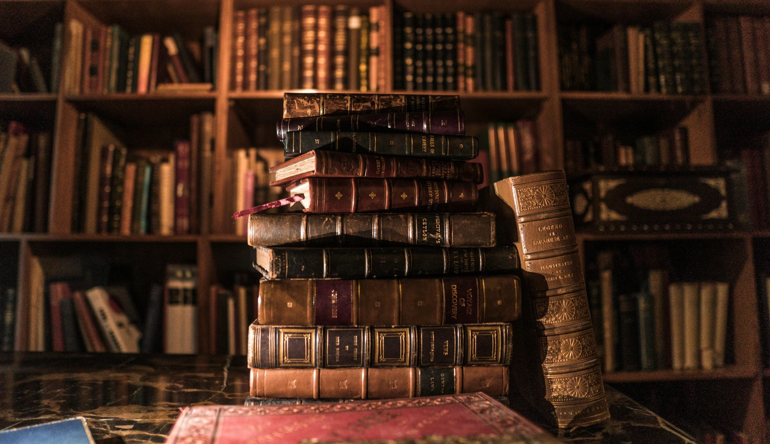 A stack of old, leather-bound books on a shelf in front of a larger bookshelf filled with many books.