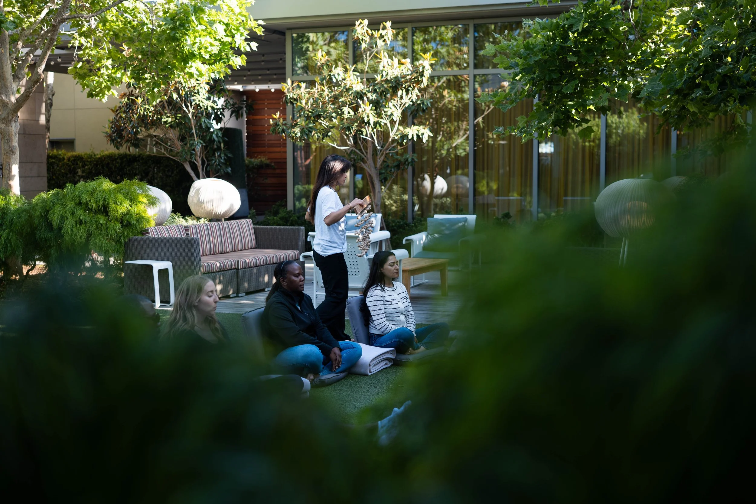 A group of women sitting cross-legged on the grass in a circle outdoors, participating in a meditation or mindfulness session, with a woman standing and reading from a notebook, surrounded by trees and modern outdoor furniture.