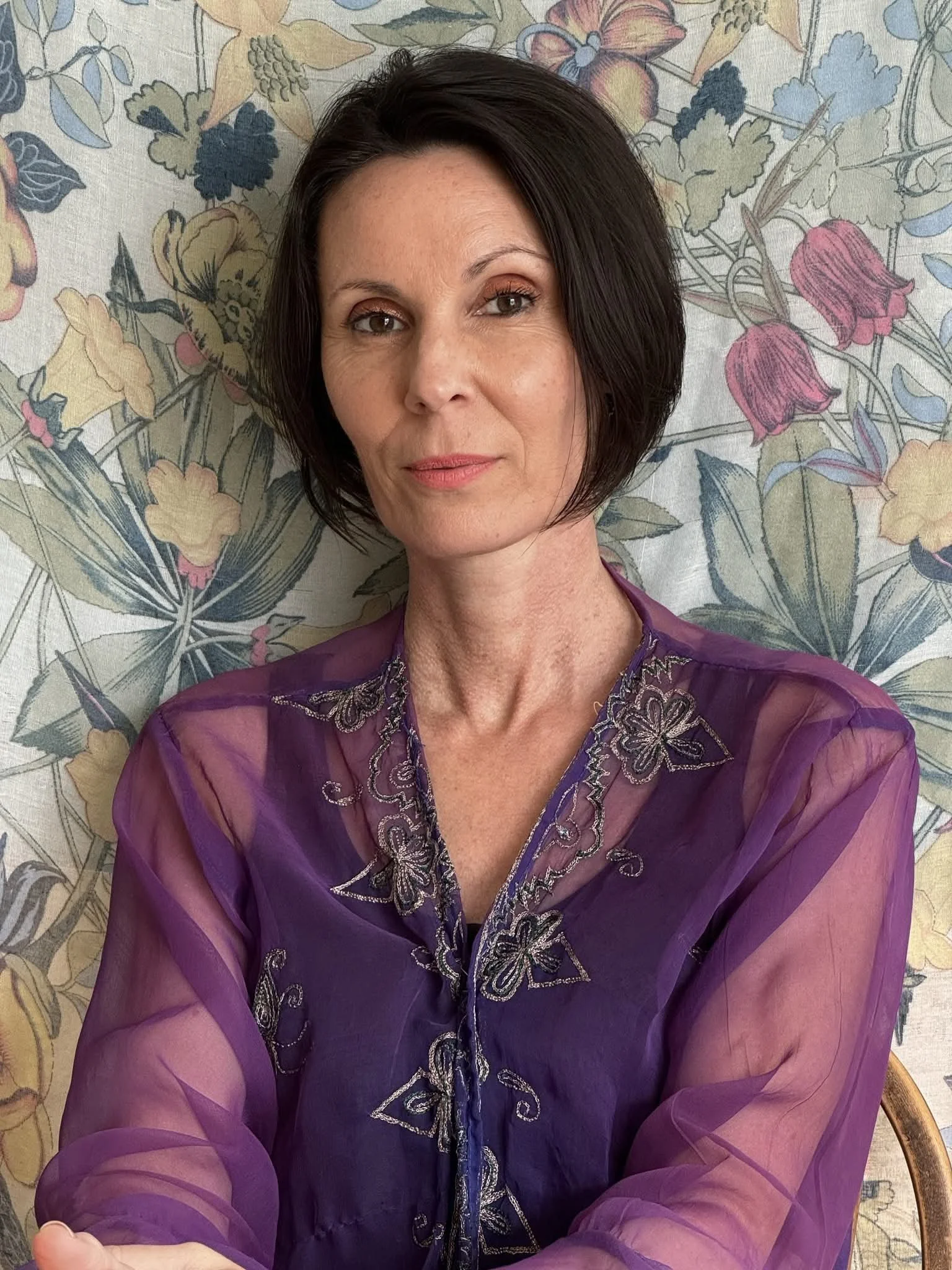 A woman with short dark hair and light skin, wearing a sheer purple embroidered blouse, sitting in front of a floral-patterned wallpaper background.