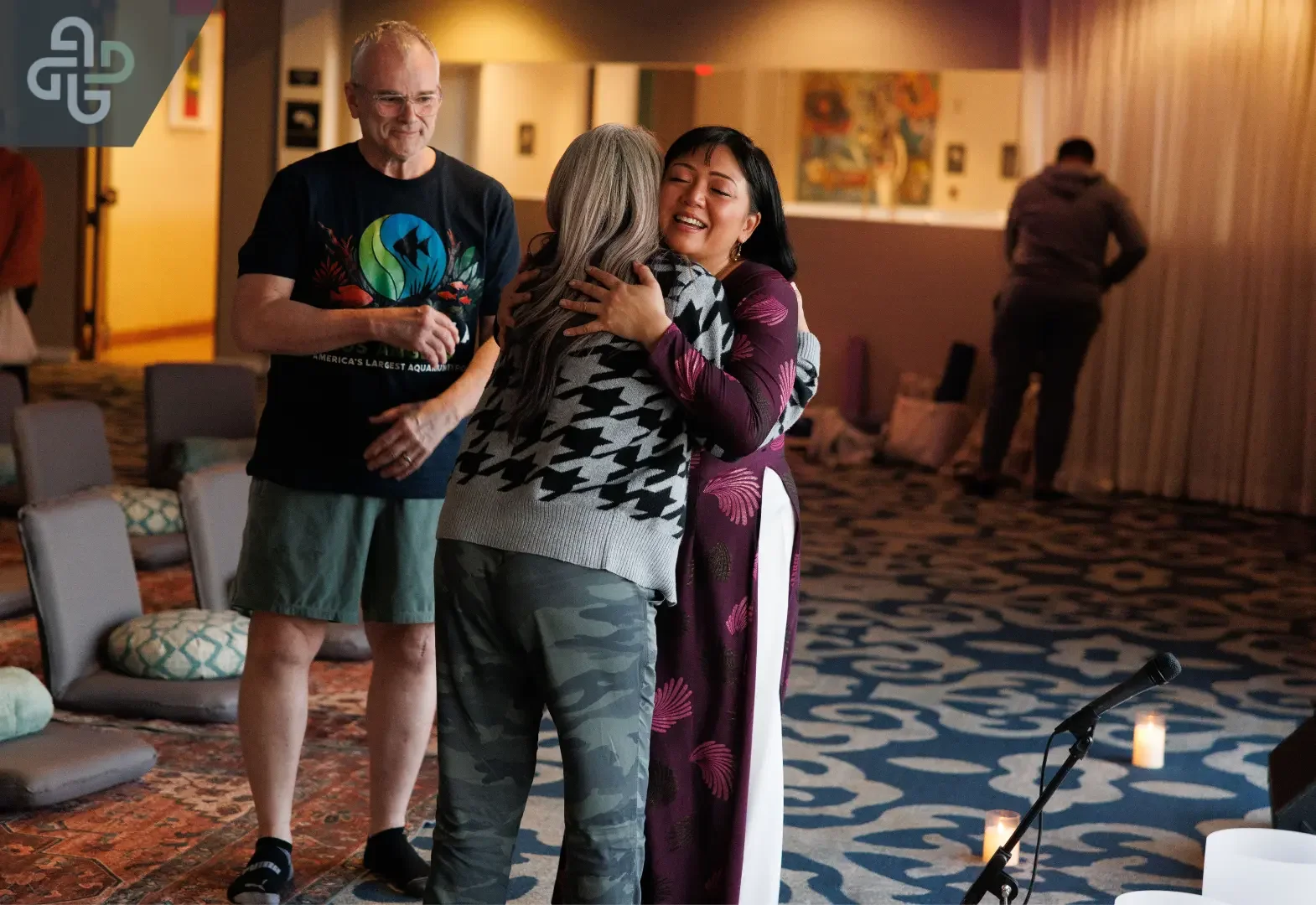 In a warmly lit community room, a smiling woman in purple hugs an older woman while a man watches, creating a welcoming, joyful scene.