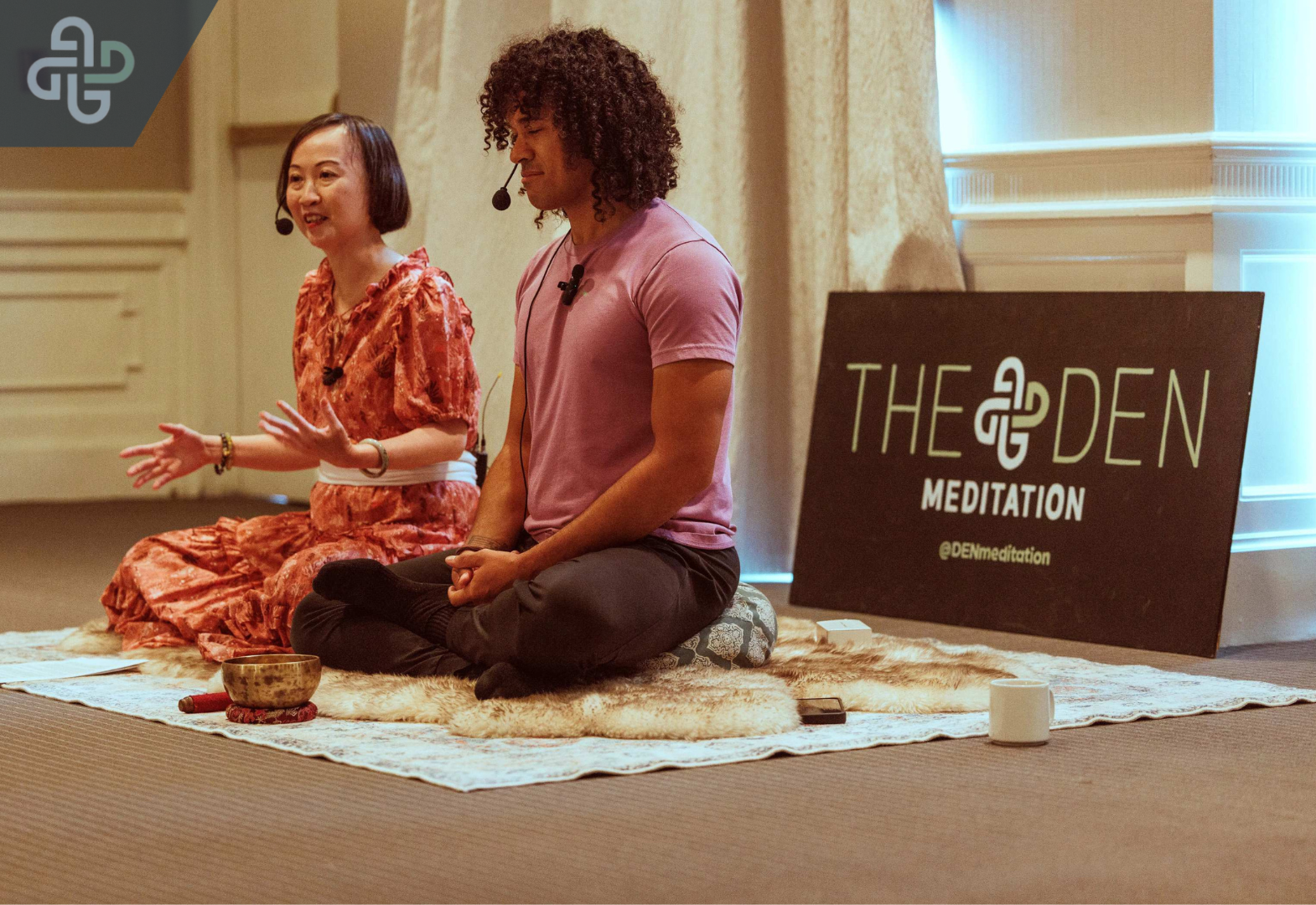 A woman speaks and gestures while a man sits in meditation on a rug. A sign nearby reads "THE DEN MEDITATION.