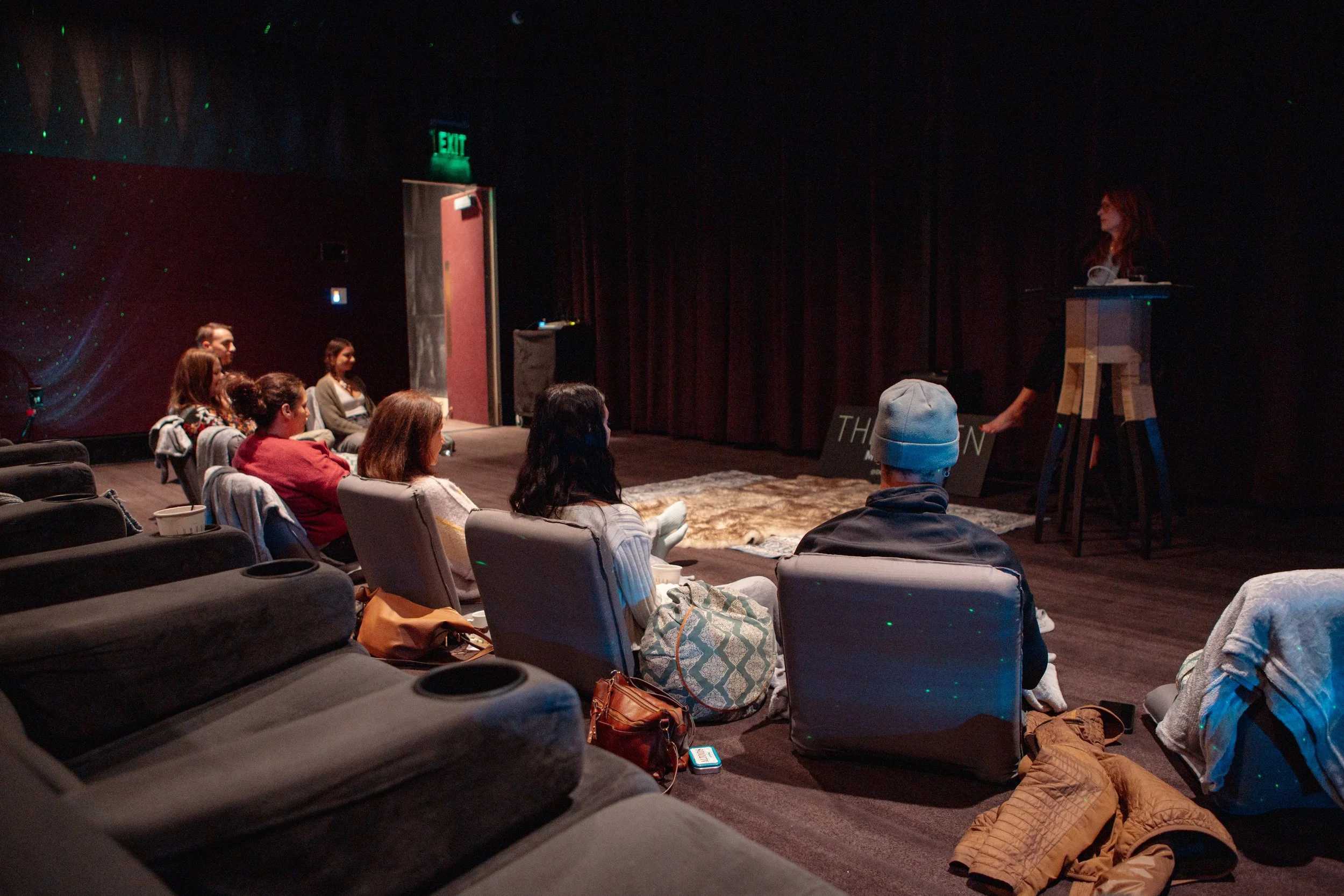 A woman standing at a podium giving a presentation to an audience in a dark theater. The audience is sitting in chairs and appears attentive, with some seated on the floor. The theater has red curtains and green exit signs.
