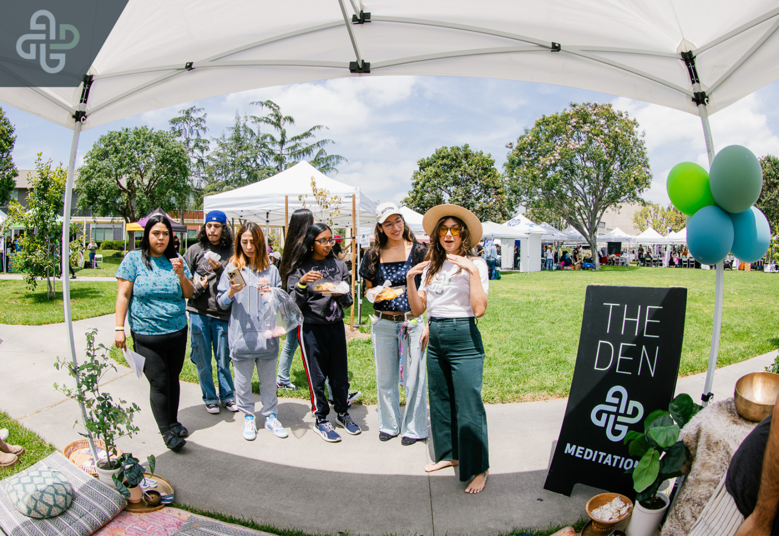 A group of people stand under a white canopy at an outdoor event. They're smiling and holding plates of food. Beside them, a sign reads "The Den Meditation." Balloons and trees enhance the festive atmosphere.