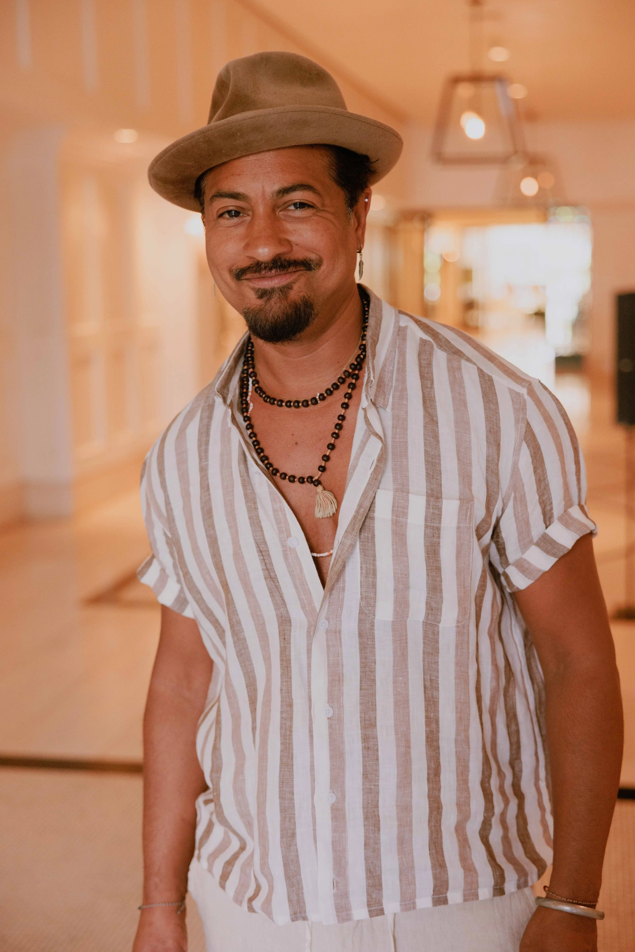 A man with a mustache and beard wearing a straw hat, striped short-sleeve shirt, and layered necklaces, standing indoors with warm lighting and wooden decor.
