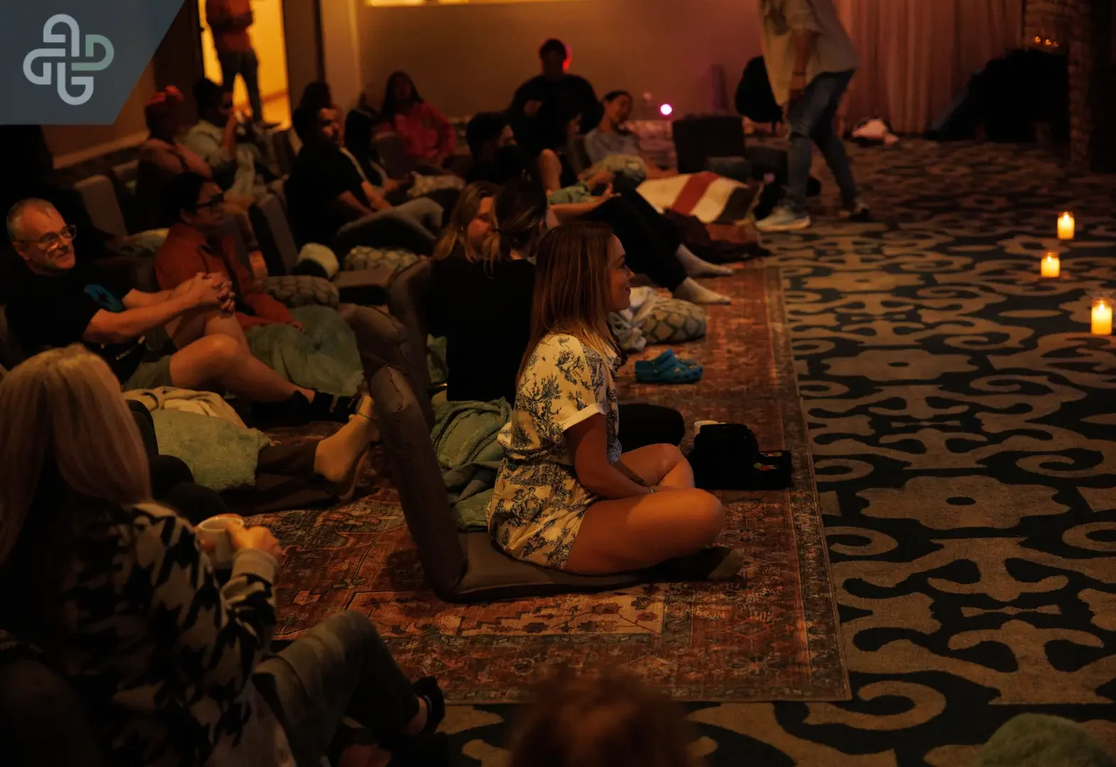 People relax on cushions and rugs in a dim, candlelit room while a woman in a floral dress sits cross-legged, creating a calm, intimate gathering.