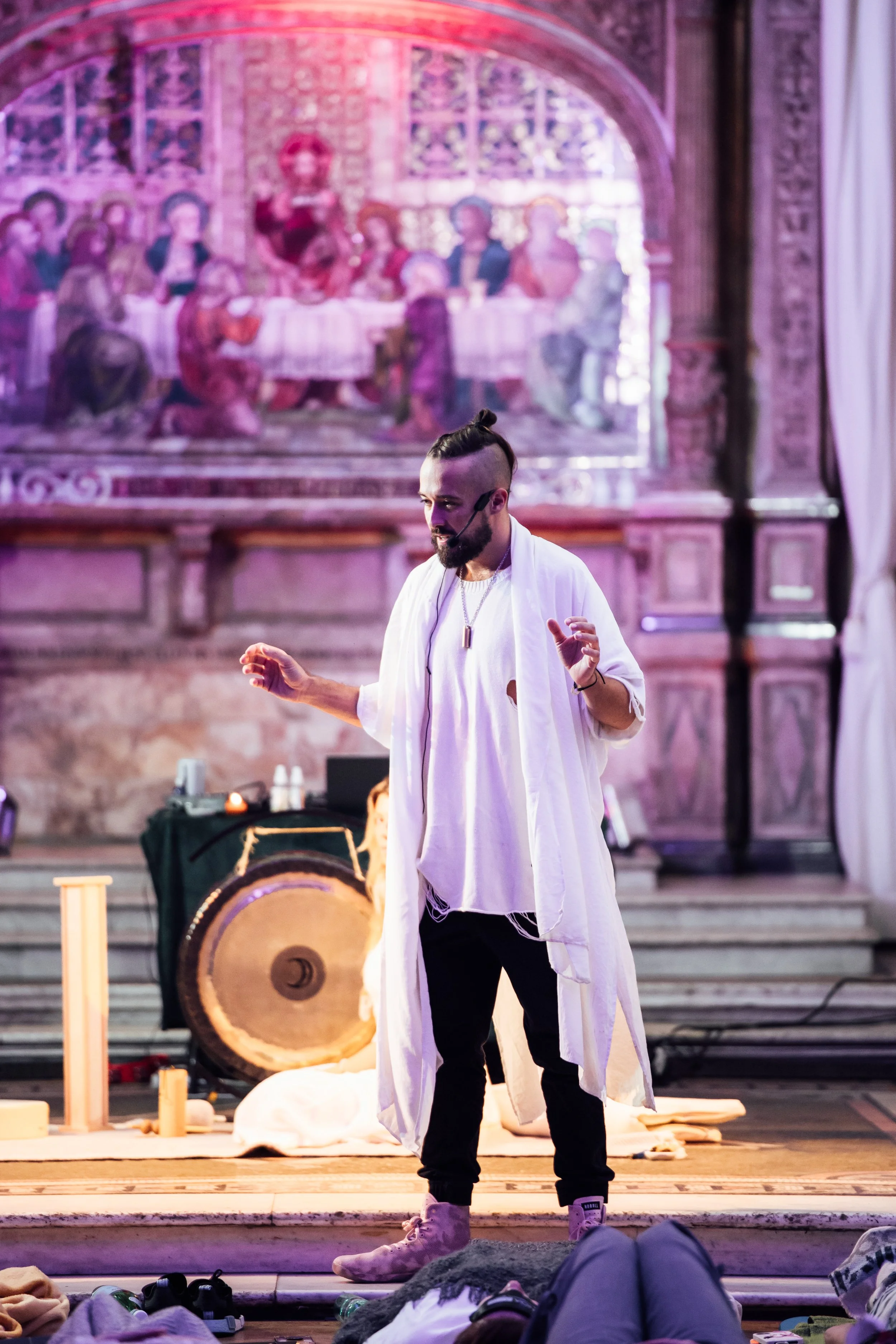 A man with a mohawk hairstyle, wearing white clothing and a headset, standing on stage. Behind him, there is a large mural or painting depicting the Last Supper. On the floor in front of him, there are musical instruments and objects, suggesting a pe