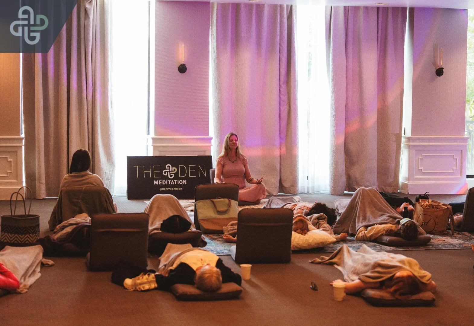 Group meditation session in a softly lit room with pink curtains; participants recline on mats while an instructor sits nearby, creating a calm mood.