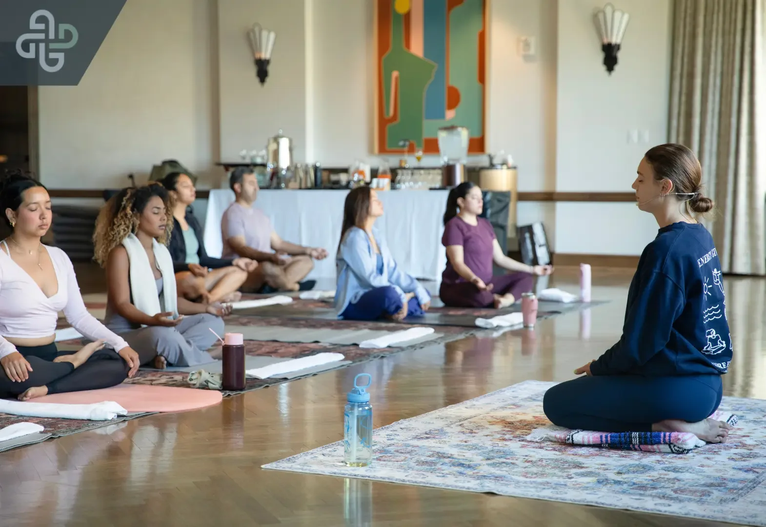 An instructor in a dark sweatshirt leads a group of people in a seated meditation session. Participants sit cross-legged on yoga mats with their eyes closed.