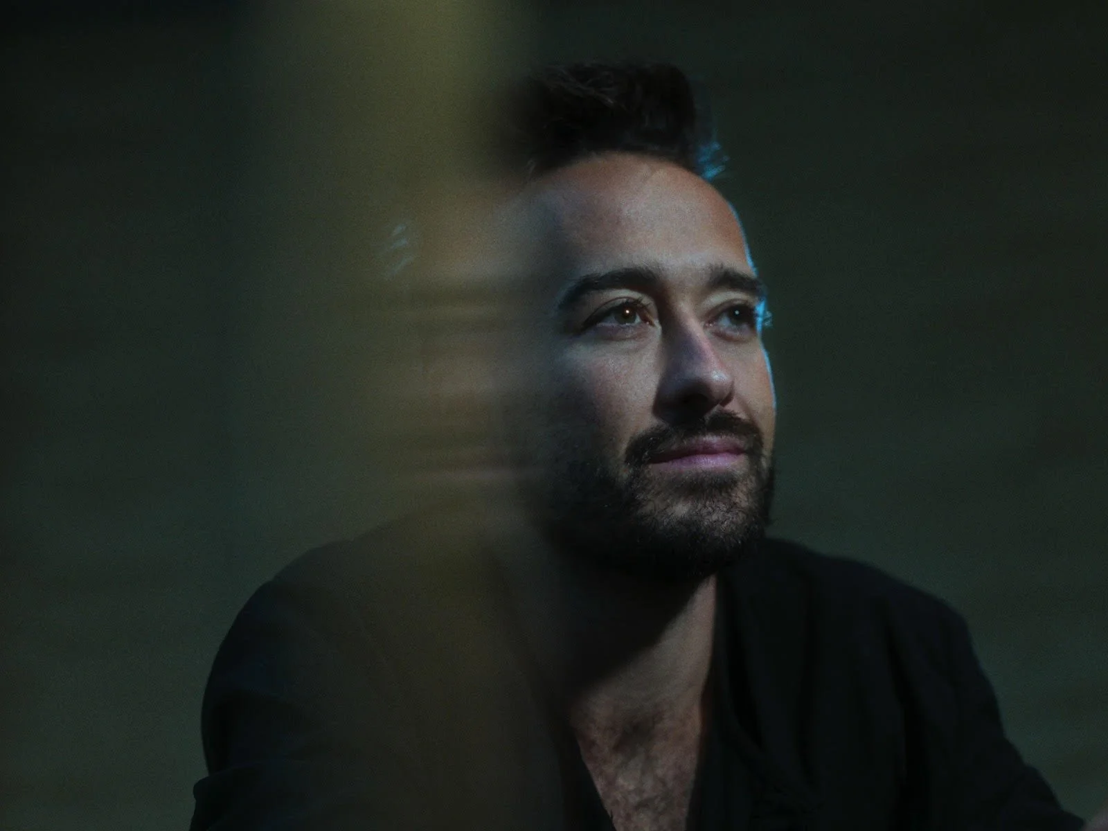 A close-up of a man's face with a beard and styled haircut, looking thoughtfully to the side against a dark background.