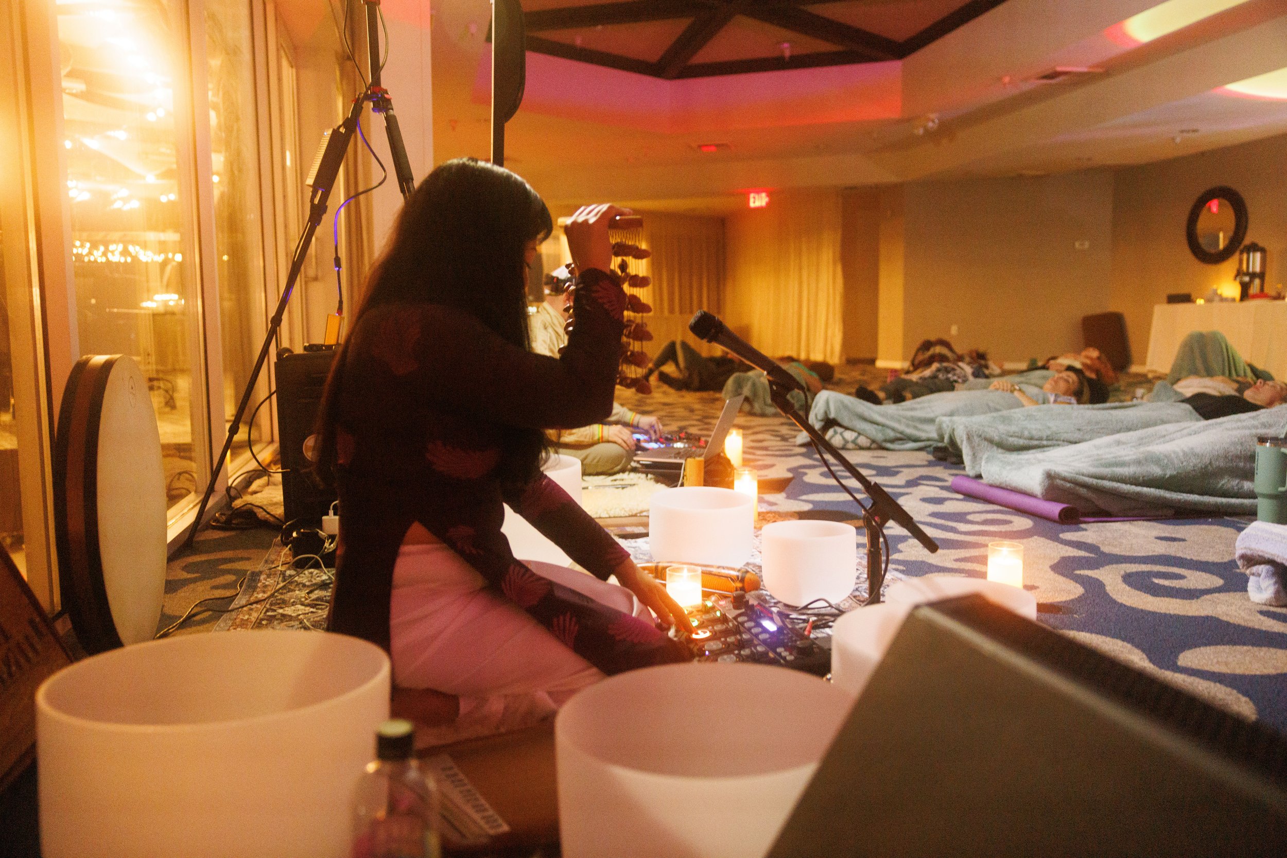 A woman with long dark hair, sitting cross-legged, operating a DJ controller, with candles around her. People are lying on blankets on the floor in a dimly lit room with warm lighting and curtains.