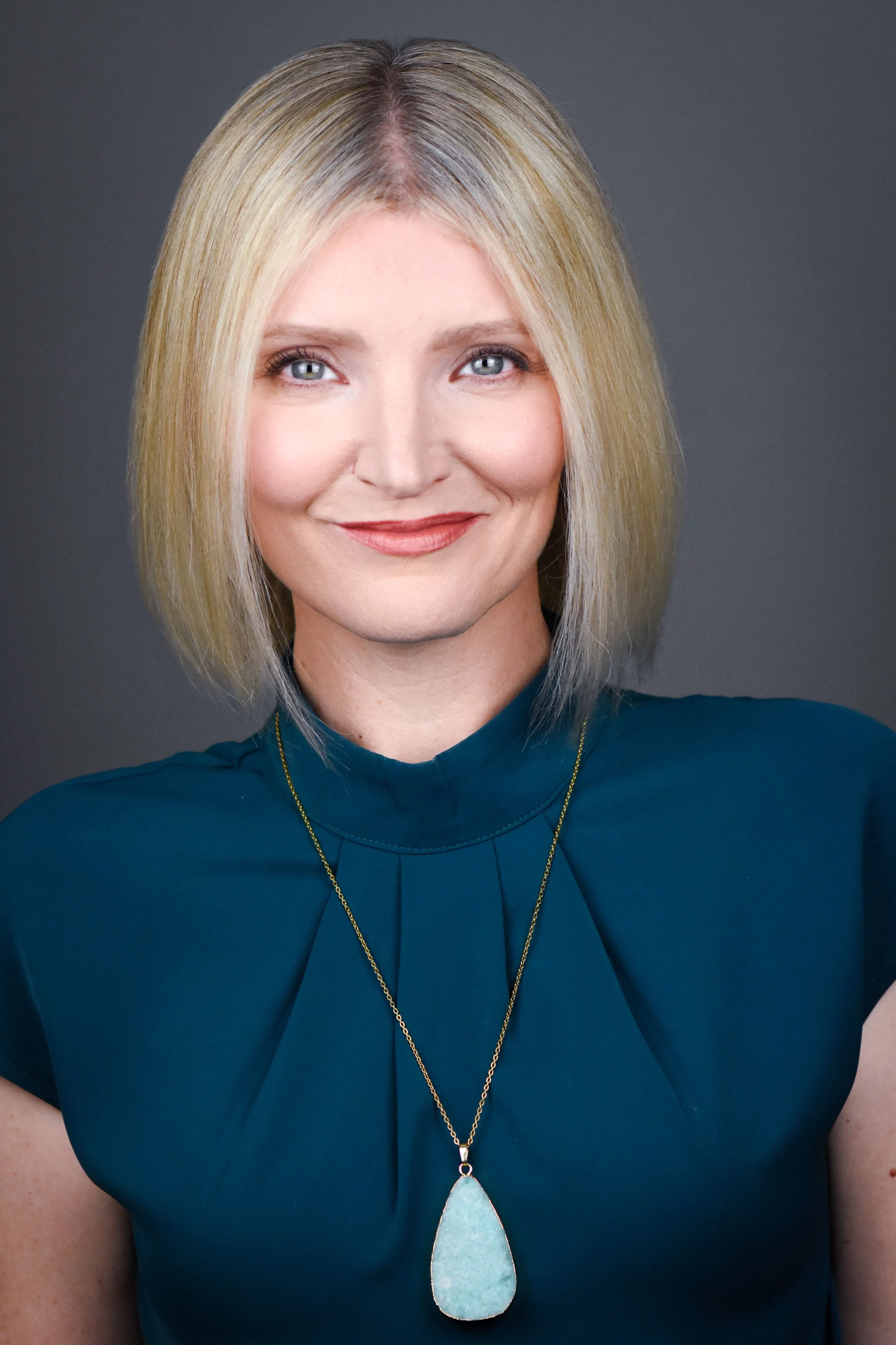 A woman with blonde hair, blue eyes, wearing a teal top, gold necklace with a large turquoise pendant, smiling.
