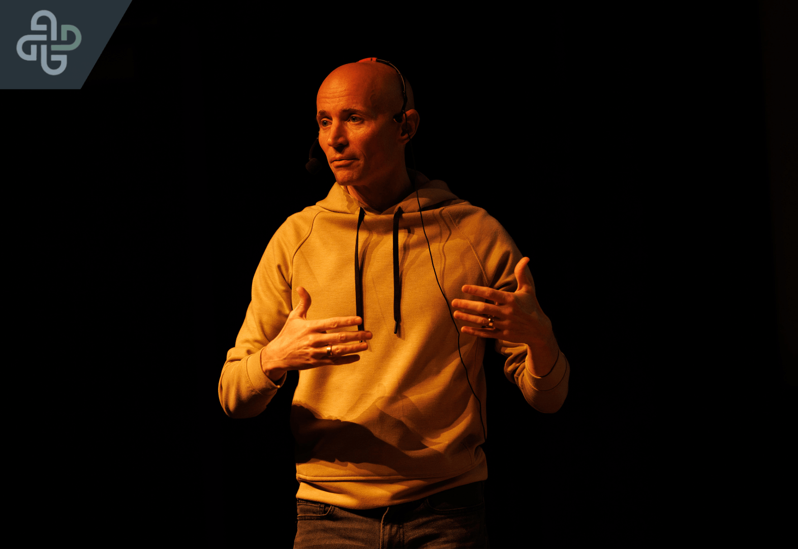 A person stands on a dark stage, wearing a headset and beige hoodie, gesturing expressively. The scene conveys focus and engagement in a presentation.
