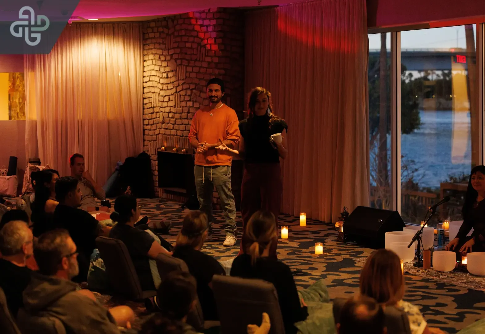 Two presenters address a seated audience in a warmly lit lounge, with candles onstage and a waterfront view through the window, creating an intimate mood.