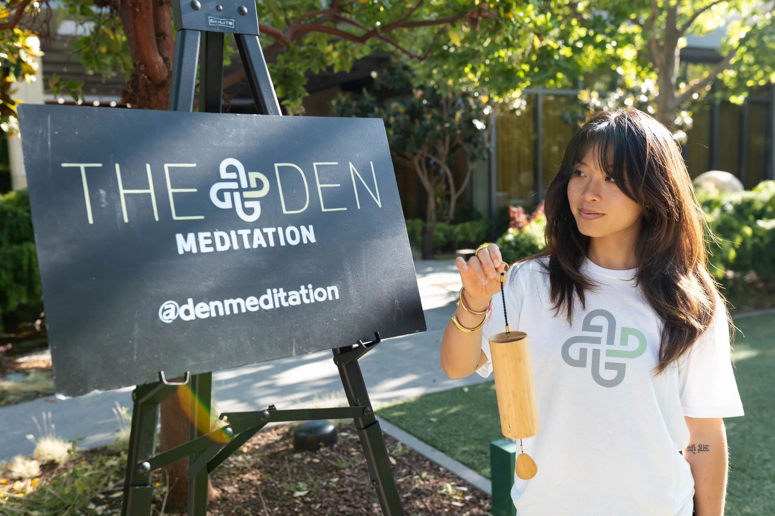 Woman standing outdoors holding a wooden singing bowl and mallet in front of a sign for The Garden Meditation, with trees and plants in the background.