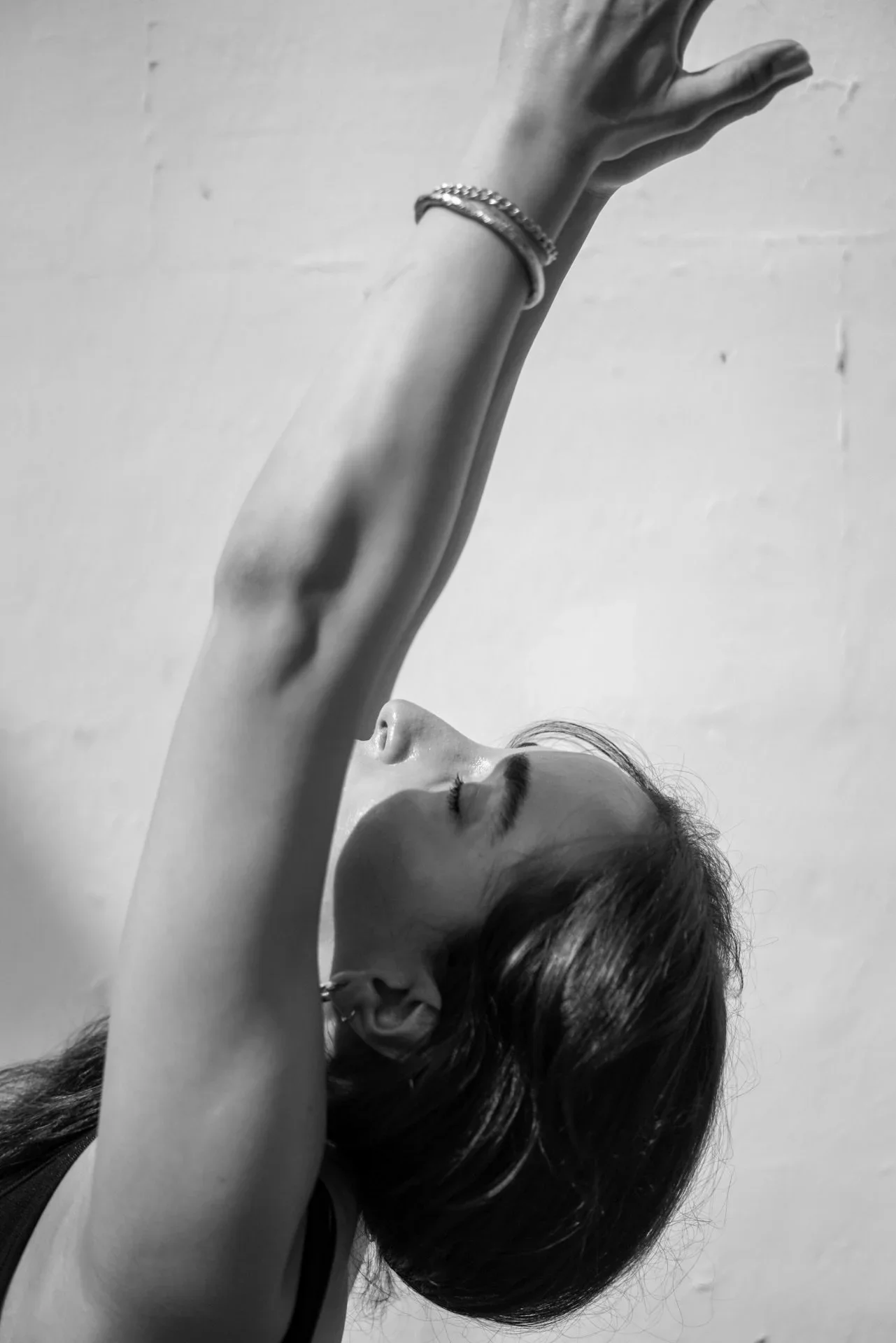 A woman with closed eyes and dark hair, wearing earrings and bracelets, stretches her arm upward against a plain wall.