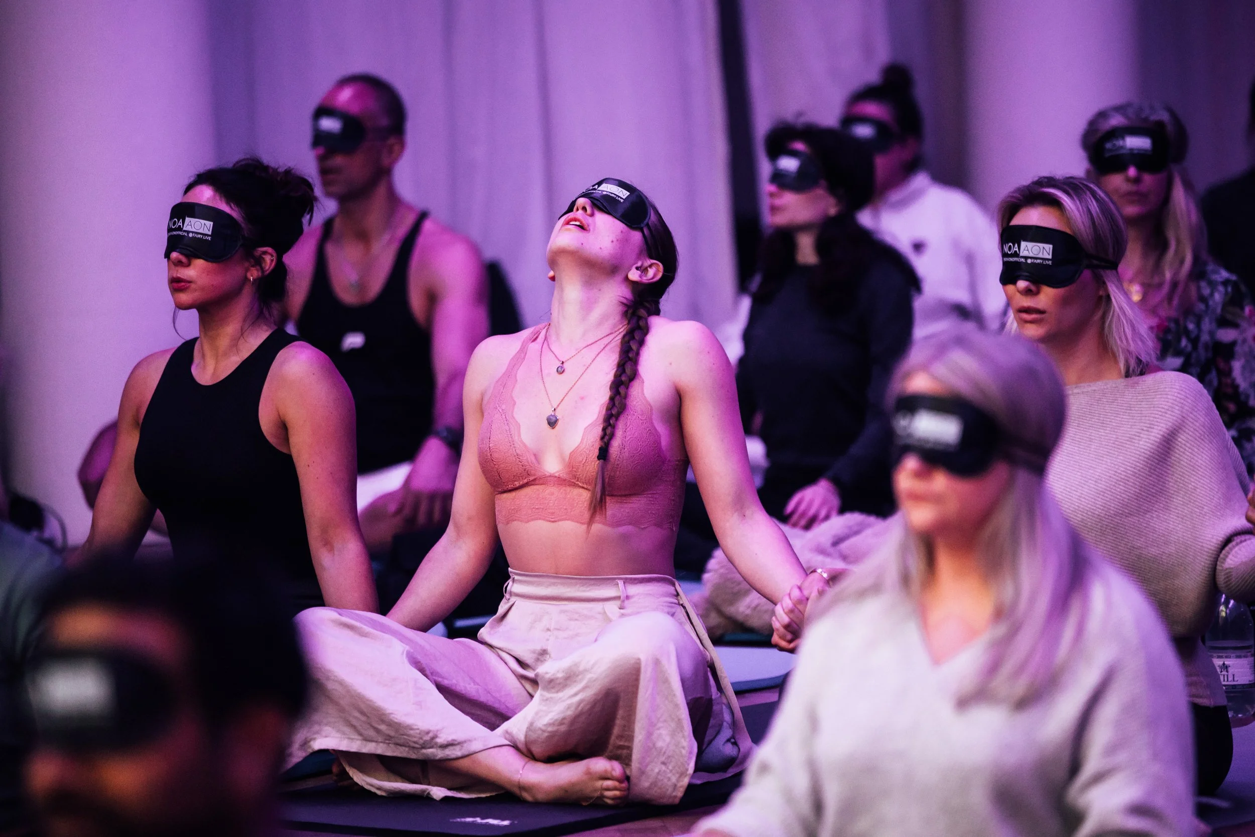 Group of people practicing meditation, wearing sleep masks, seated on the floor with legs crossed, in a dimly lit room.