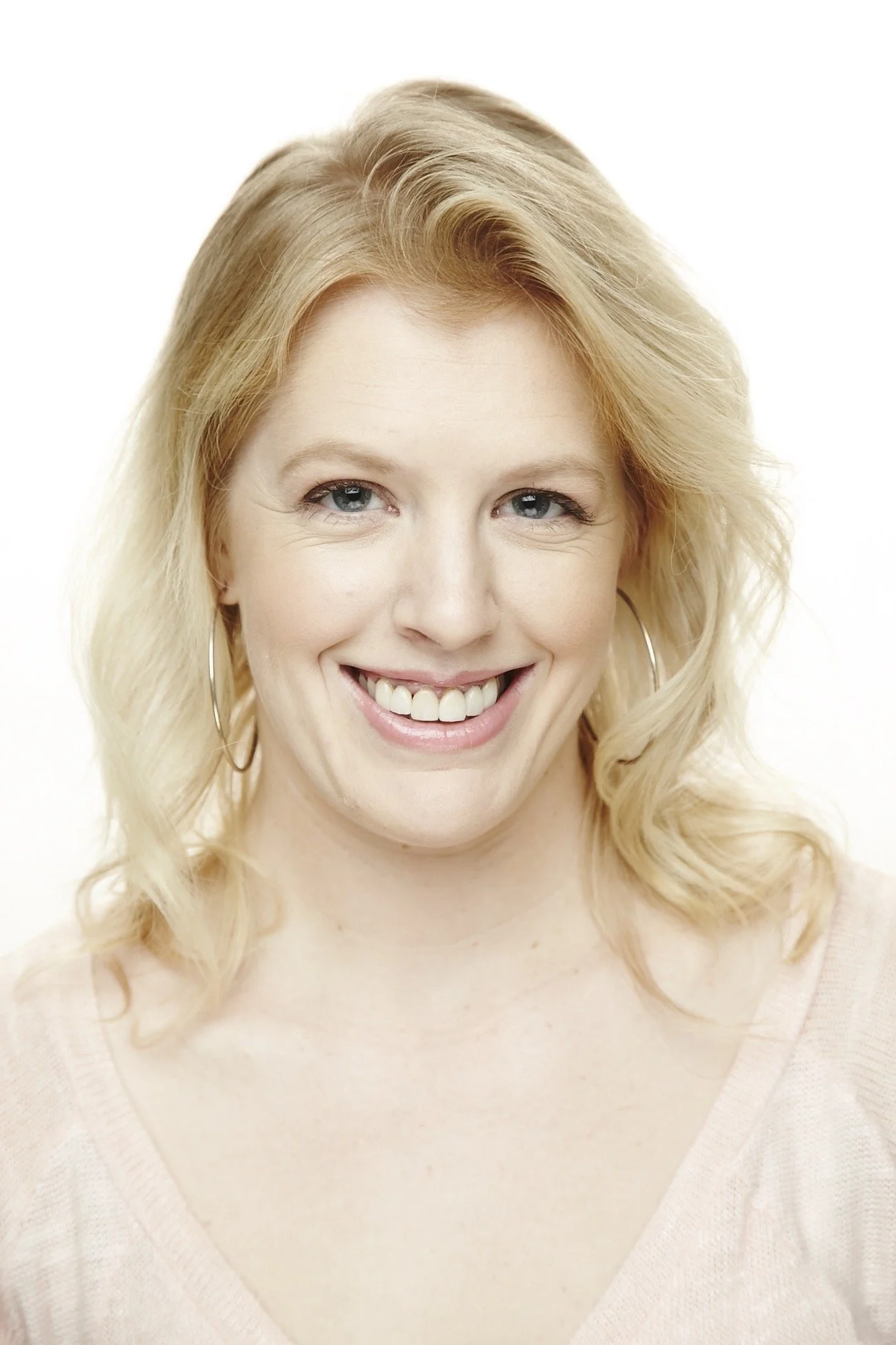 A woman with shoulder-length blonde hair, blue eyes, and a bright smile, wearing a light-colored top, against a plain white background.