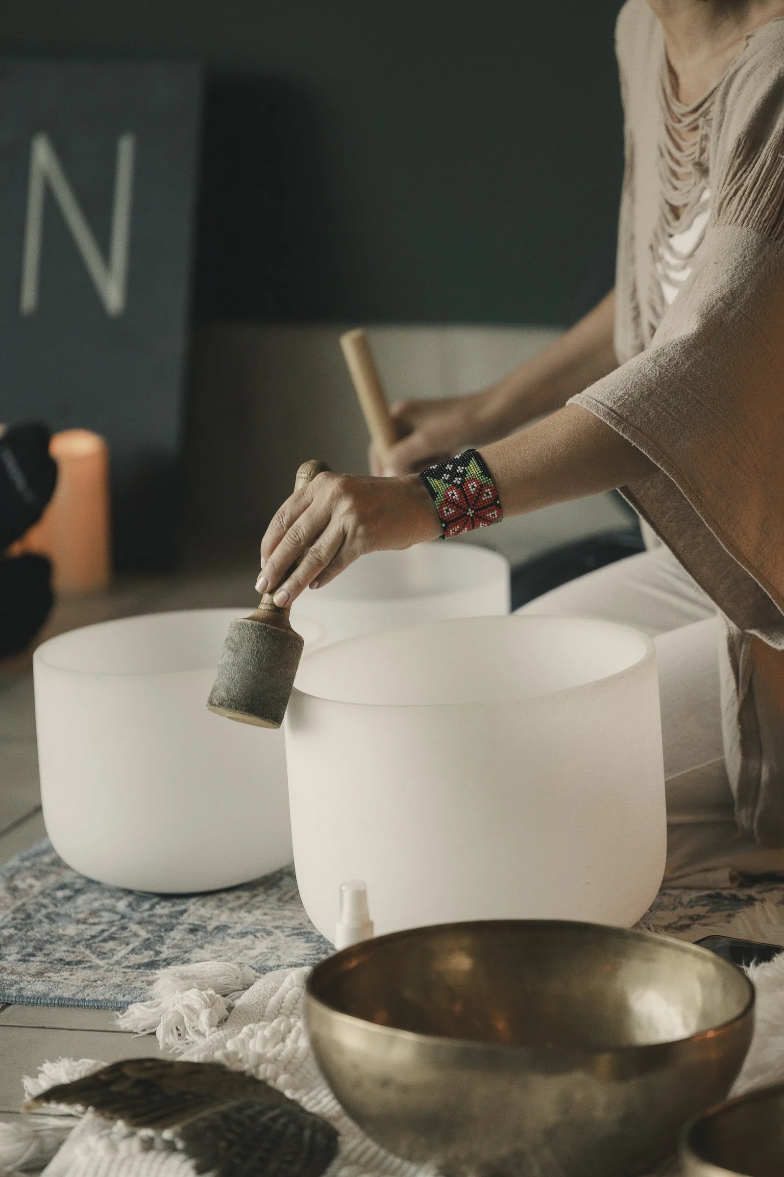 Person playing crystal singing bowls, holding a mallet, with a colorful beaded bracelet, on a table with other singing bowls and a textured cloth.