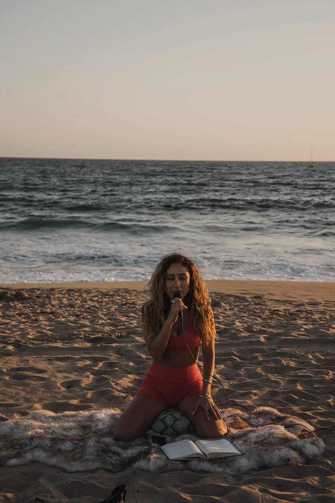 Woman with curly hair singing into a microphone while kneeling on a fur rug on the beach during sunset, with an open book in front of her.