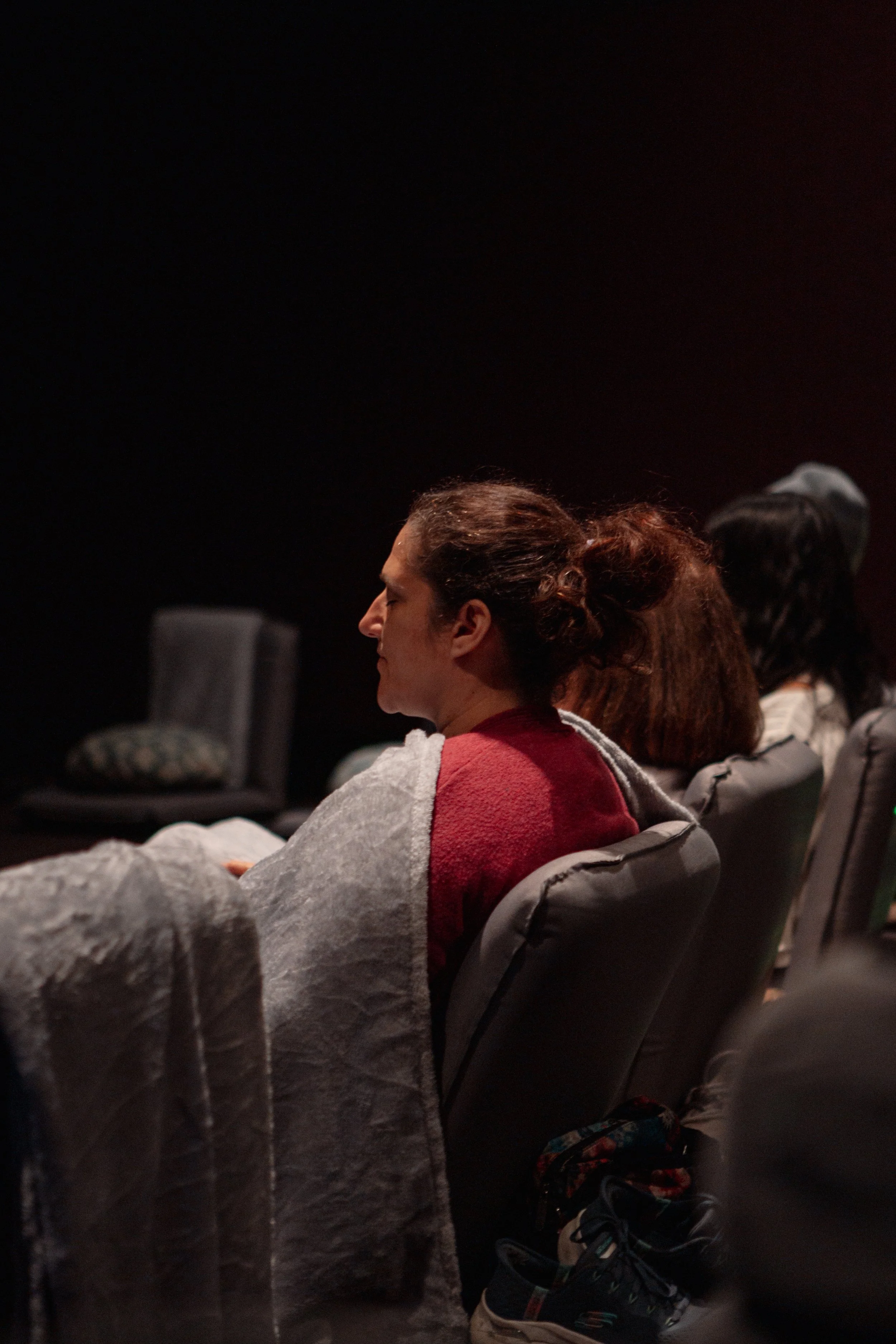 People seated and meditating in a dark room, faces turned to the side with closed eyes.