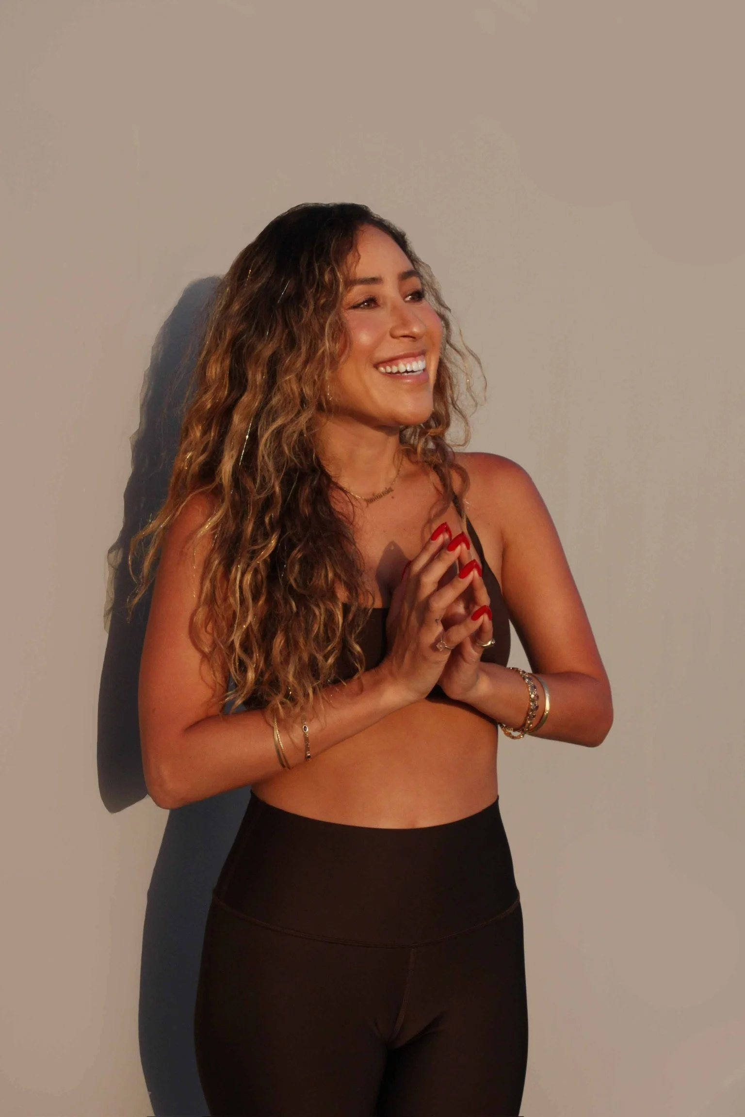A smiling woman with curly hair And tan skin, wearing a black sports bra and high-waisted leggings, standing against a plain wall with her hands clasped in front of her chest.