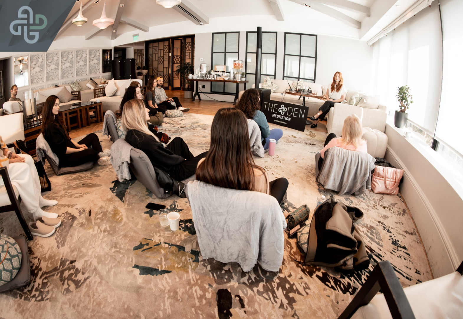 A group of people sit on cushions in a bright room, engaged in a meditation session. A sign reads "The Den Meditation," creating a calm atmosphere.
