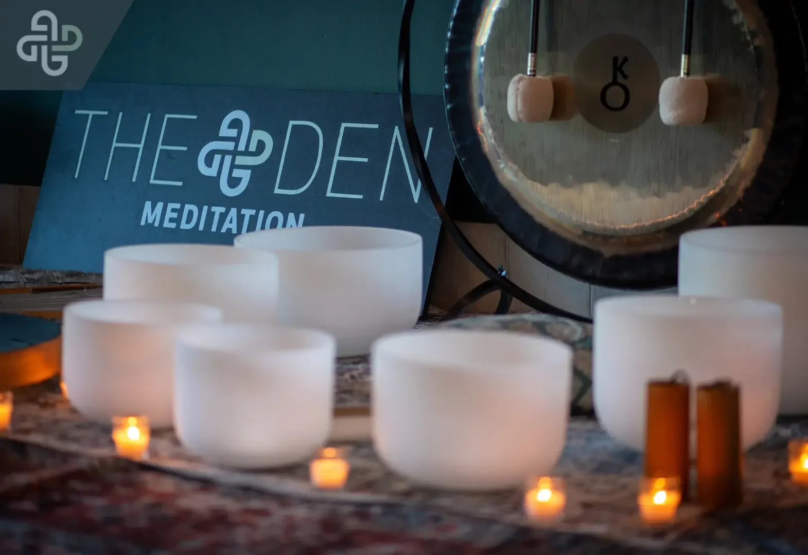 White crystal singing bowls, candles, and a large gong create a serene meditation setup, with a “The Den Meditation” sign in the background.