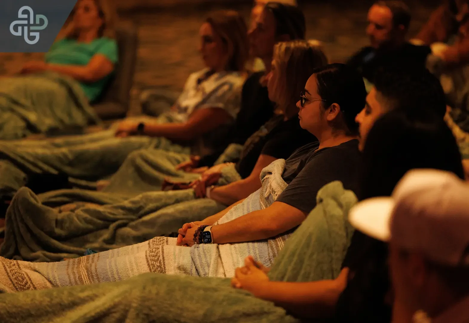 Adults sit quietly in a row, wrapped in blankets in a dim, warmly lit room; their attentive posture creates a calm, reflective group atmosphere.