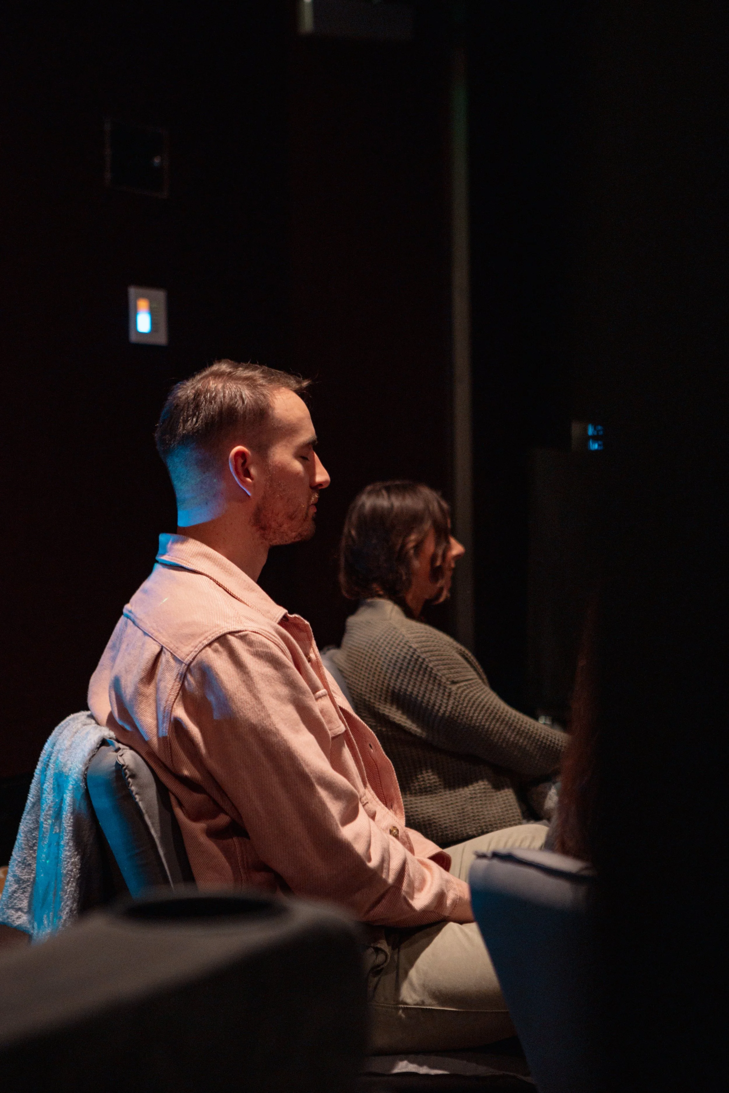 Two people sitting cross-legged on chairs with eyes closed in a dark room, possibly meditating or attending a meditation class.