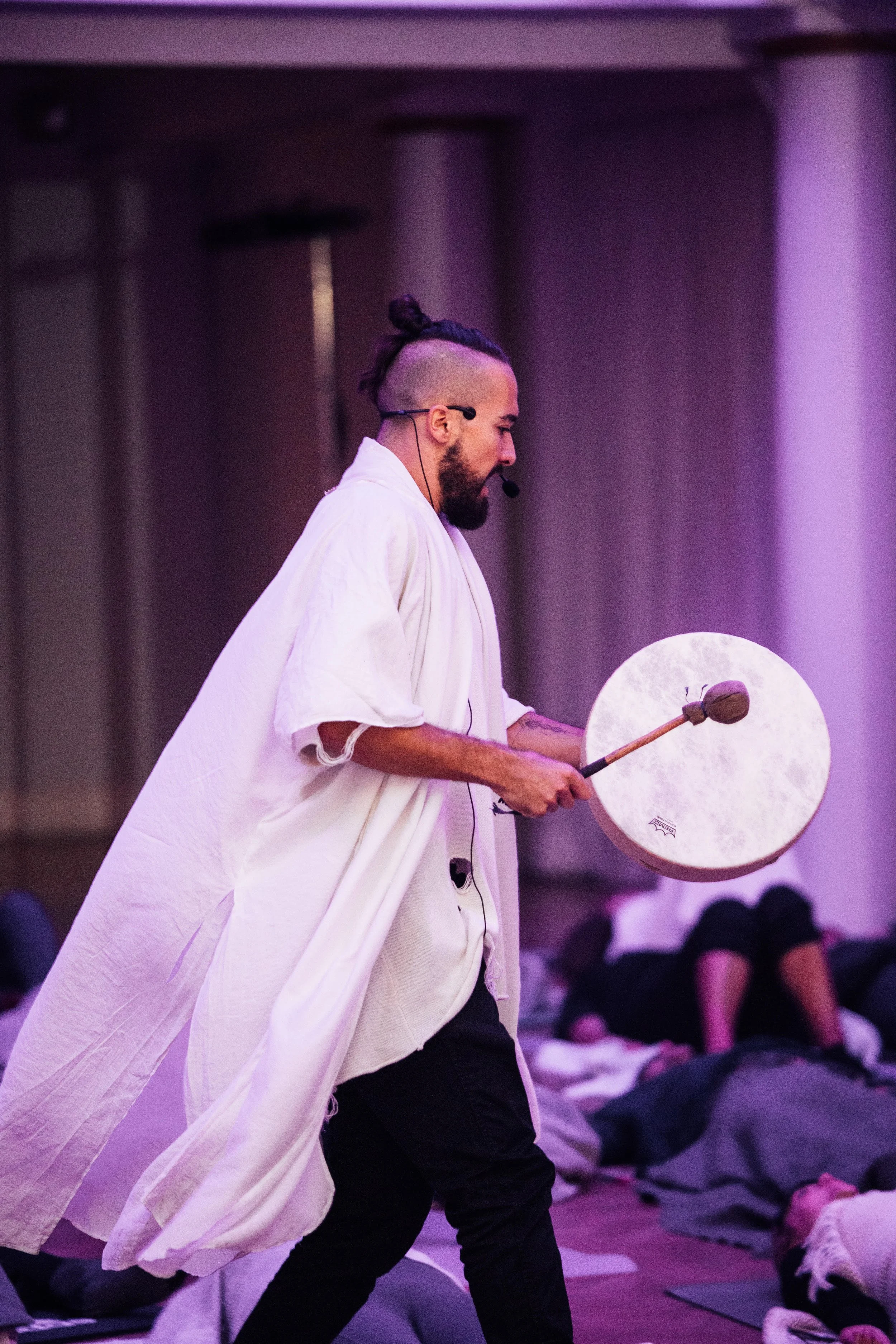 Man with a beard, dressed in a white robe, holding a gong and a mallet, leading a meditation session with several people lying on mats in the background.