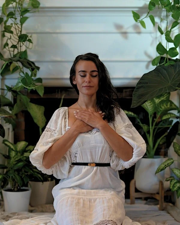 Woman with dark hair, sitting cross-legged with eyes closed and hands on chest, surrounded by green houseplants.