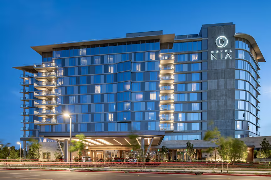 Exterior of a modern hotel building with glass windows, illuminated balconies, and a sign reading 'Hotel NIA' during dusk.