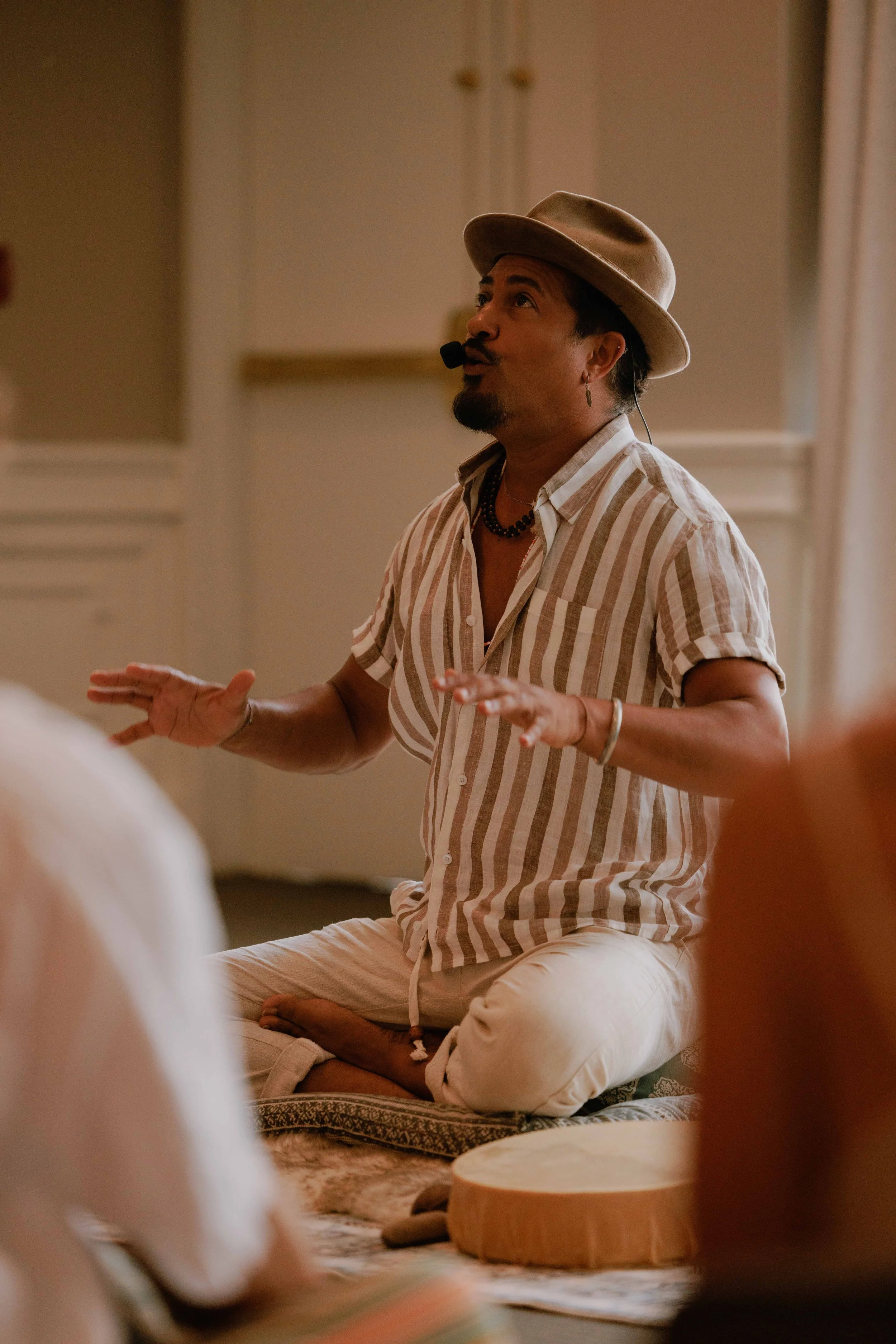 A man wearing a beige hat, striped shirt, and white pants is sitting cross-legged and speaking with hands raised, in a room with beige walls.