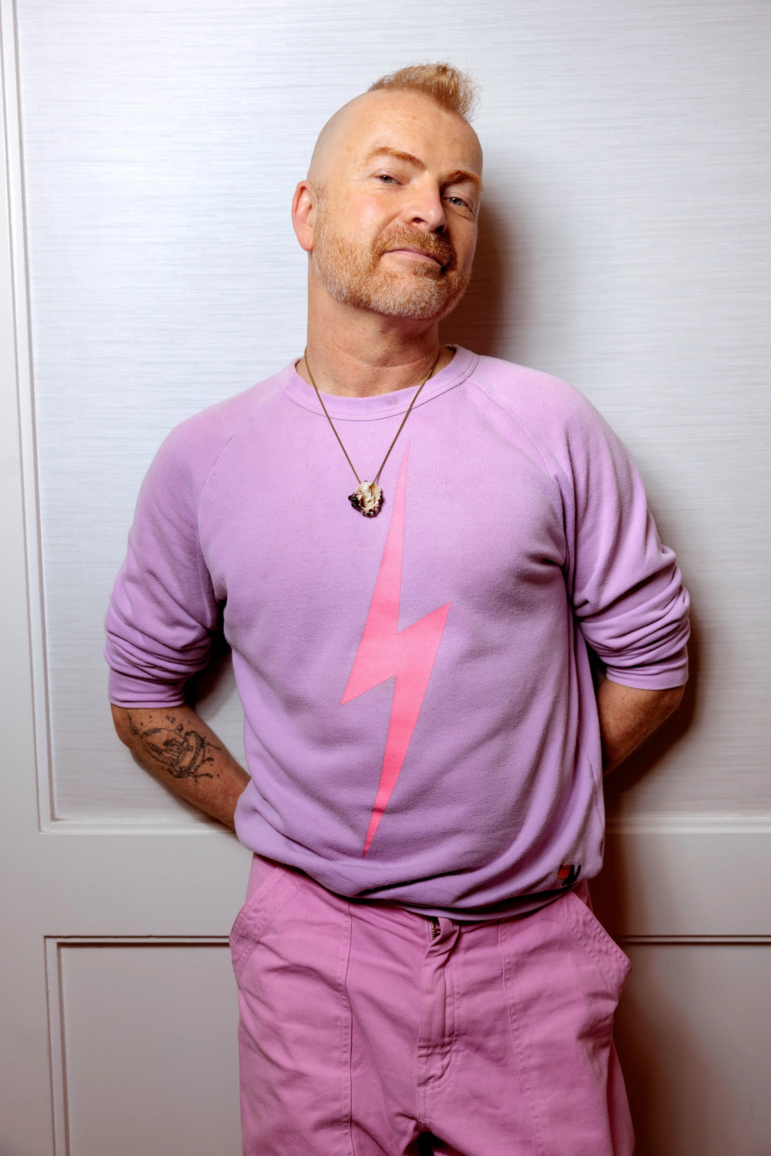 Man with a beard and mustache leaning against a wall, wearing a pink sweatshirt with a lightning bolt, pink pants, a necklace, and tattoos on his arm.