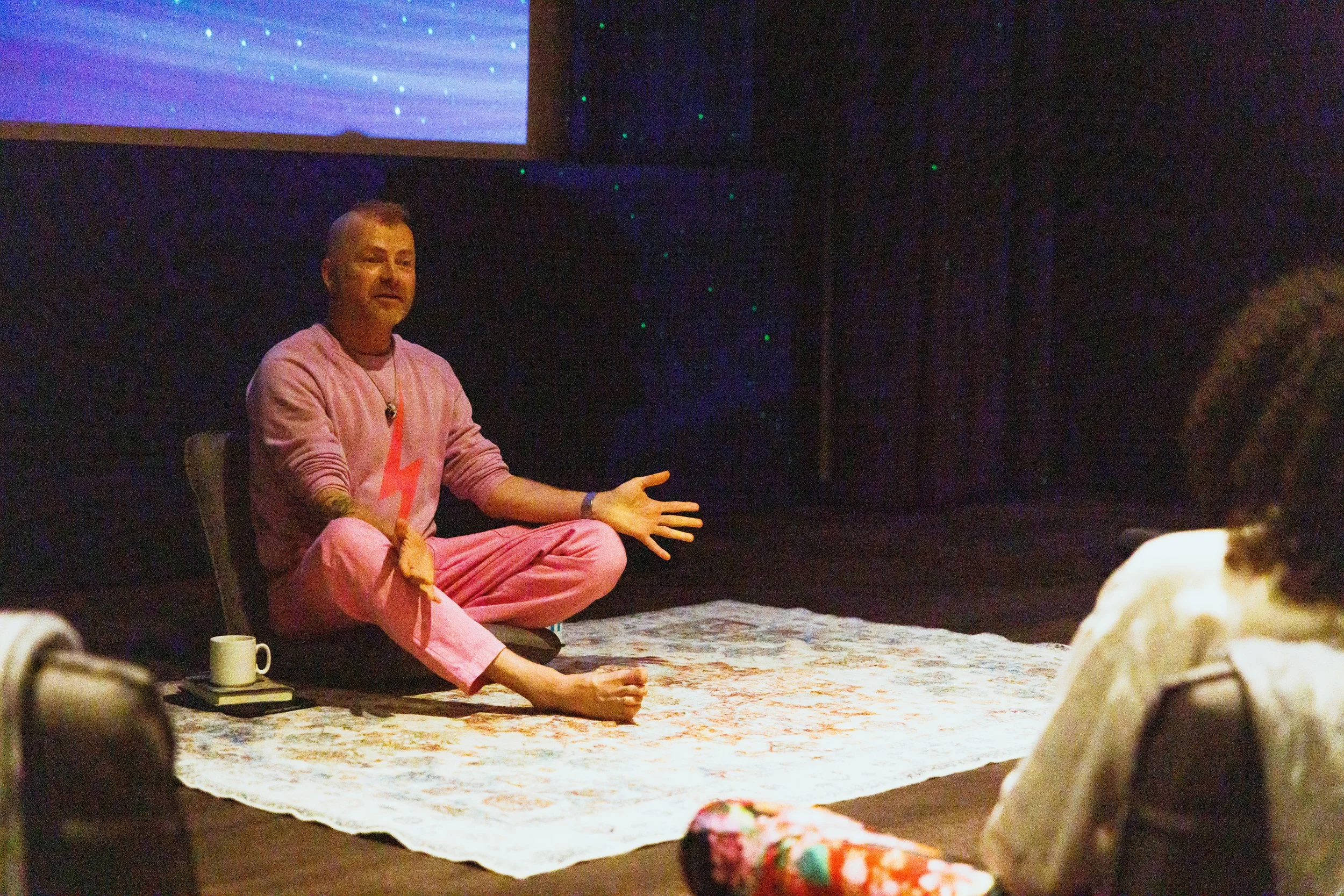 A man dressed in pink, sitting cross-legged on a rug, gesturing with his hand while talking to a group in a dark room with a projector screen in the background.