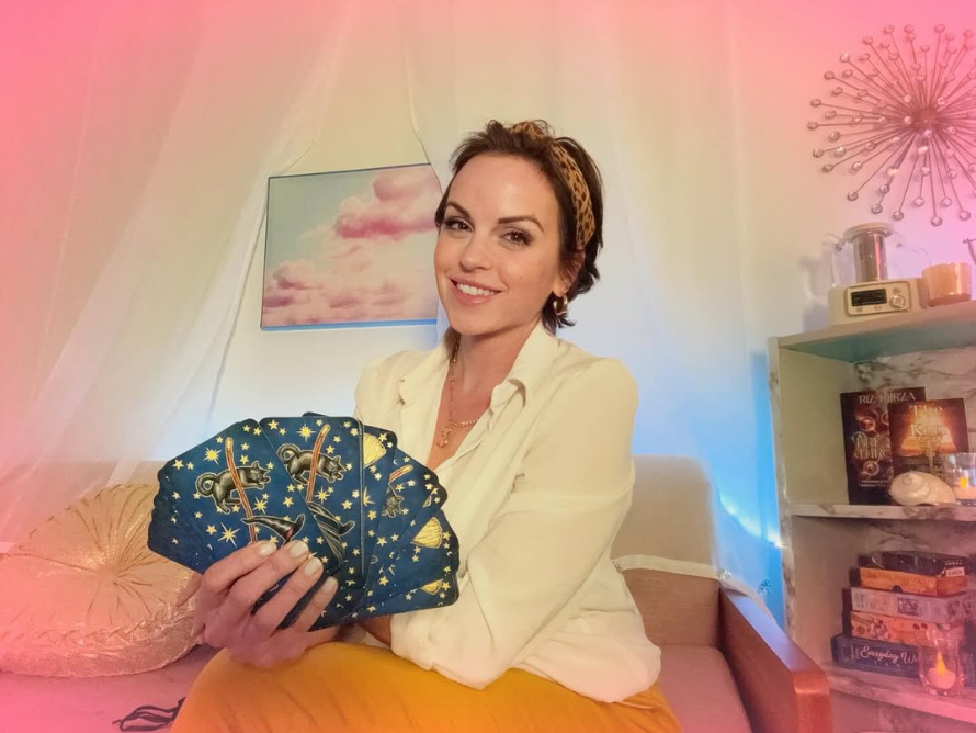 A woman with short brown hair, wearing a yellow headband and a cream-colored shirt, sitting on a beige couch holding a fan of tarot cards with starry, celestial designs, in a room with colorful lighting.