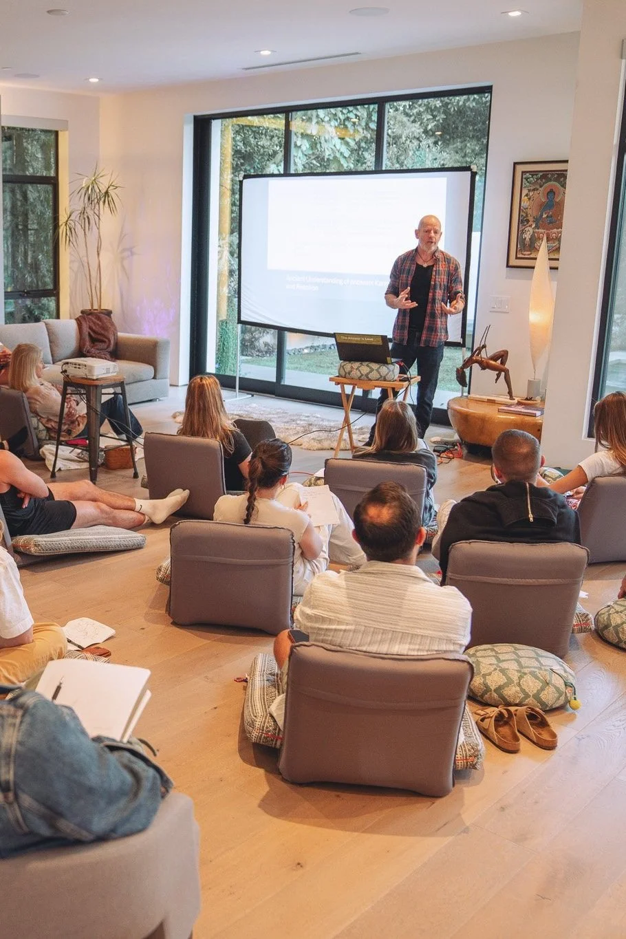 A man giving a presentation in a cozy, modern living room with large windows and a view of trees outside. The audience sits on cushions and chairs on the floor, taking notes. There is a projector screen behind the speaker, a laptop on a small table, and decorative items like a sculpture and artwork.