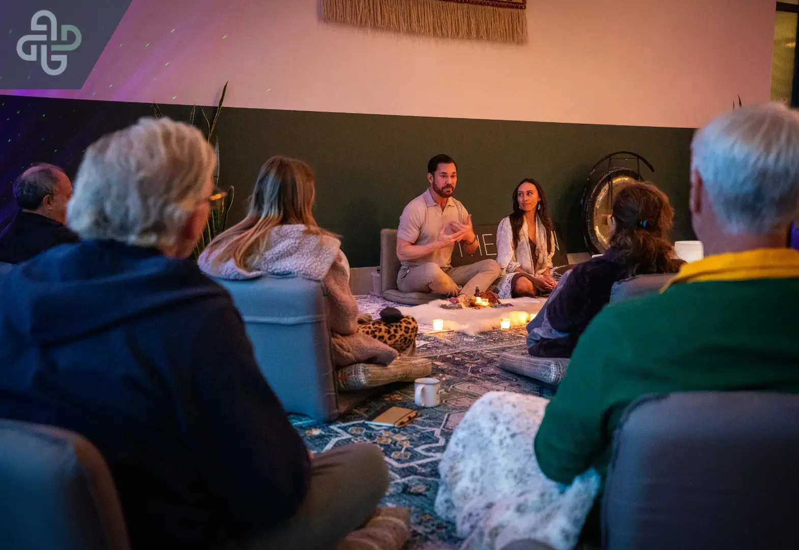 Small group gathers in a warmly lit room for a seated discussion or meditation circle, creating an intimate, calm, reflective atmosphere.