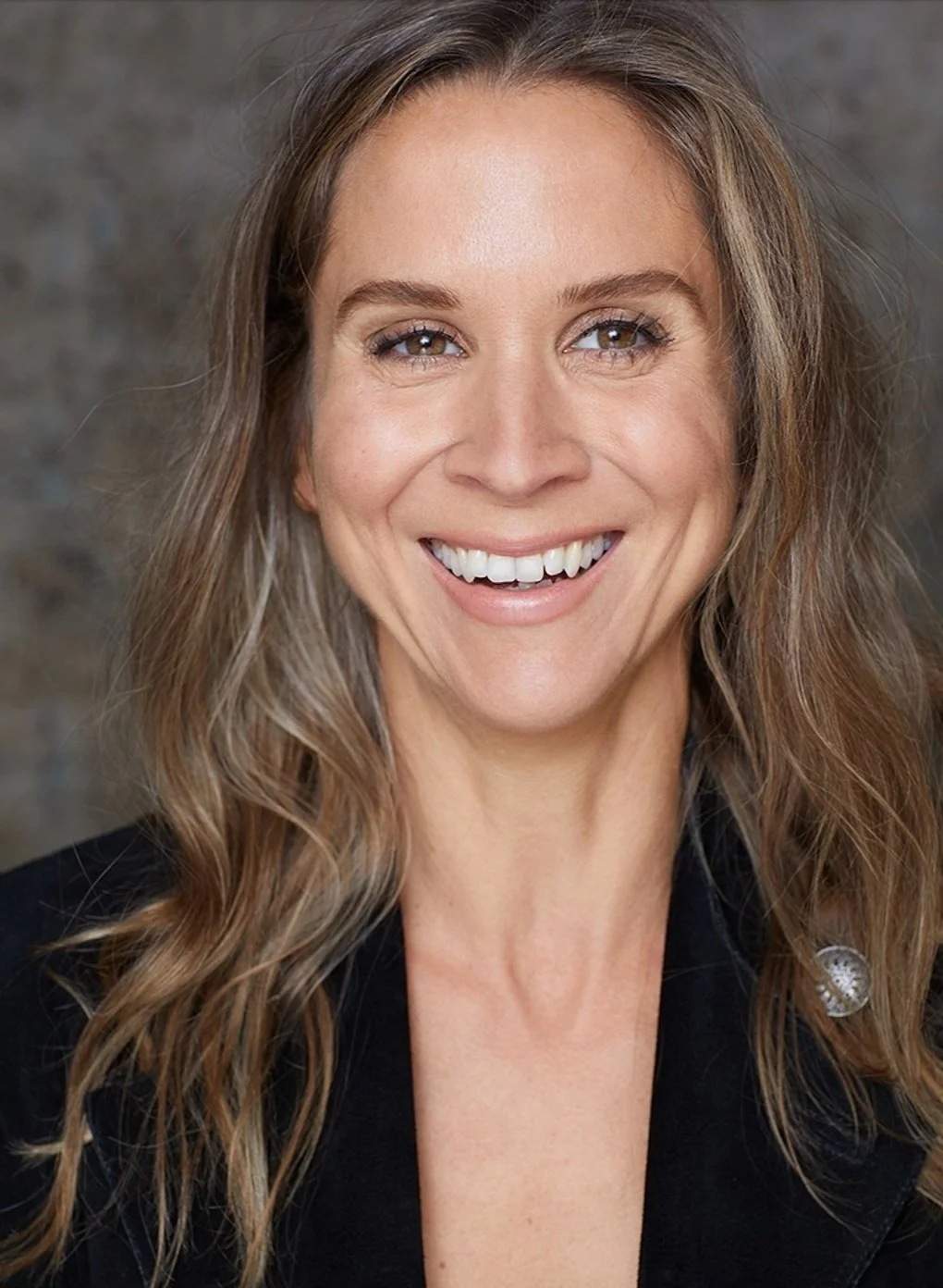 A smiling woman with long, wavy light brown hair, wearing a black top, against a blurred gray background.