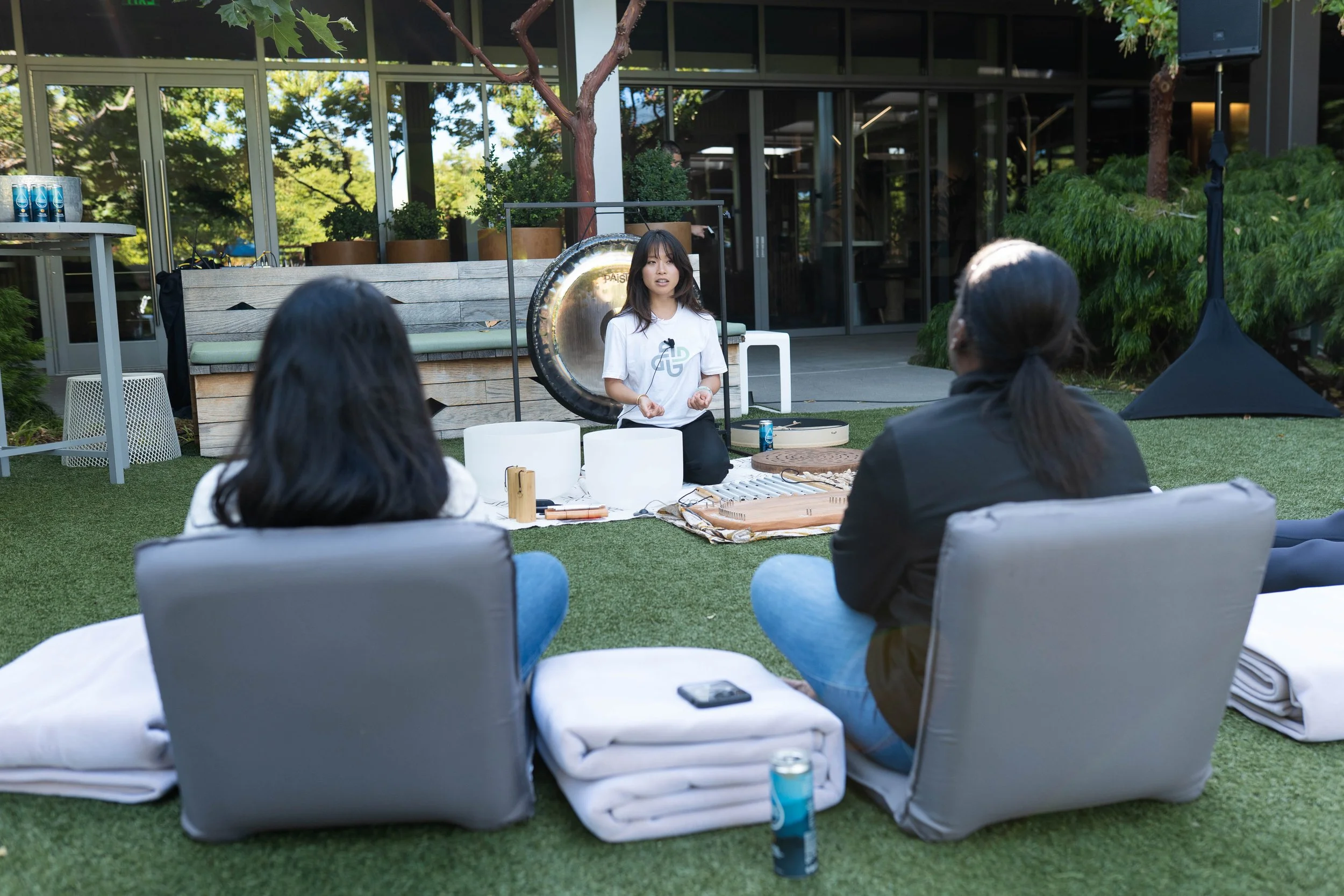 A woman is standing in front of a small audience outside, surrounded by musical instruments, including a gong and singing bowls, participating in a wellness or meditation session, with a building and trees in the background.