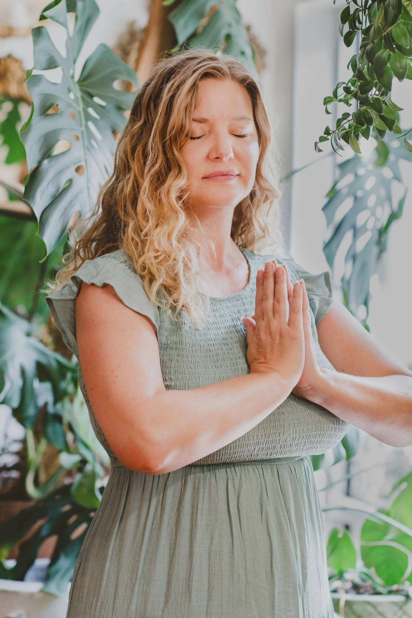 A woman practicing yoga with her eyes closed, hands pressed together in prayer position, surrounded by green plants.