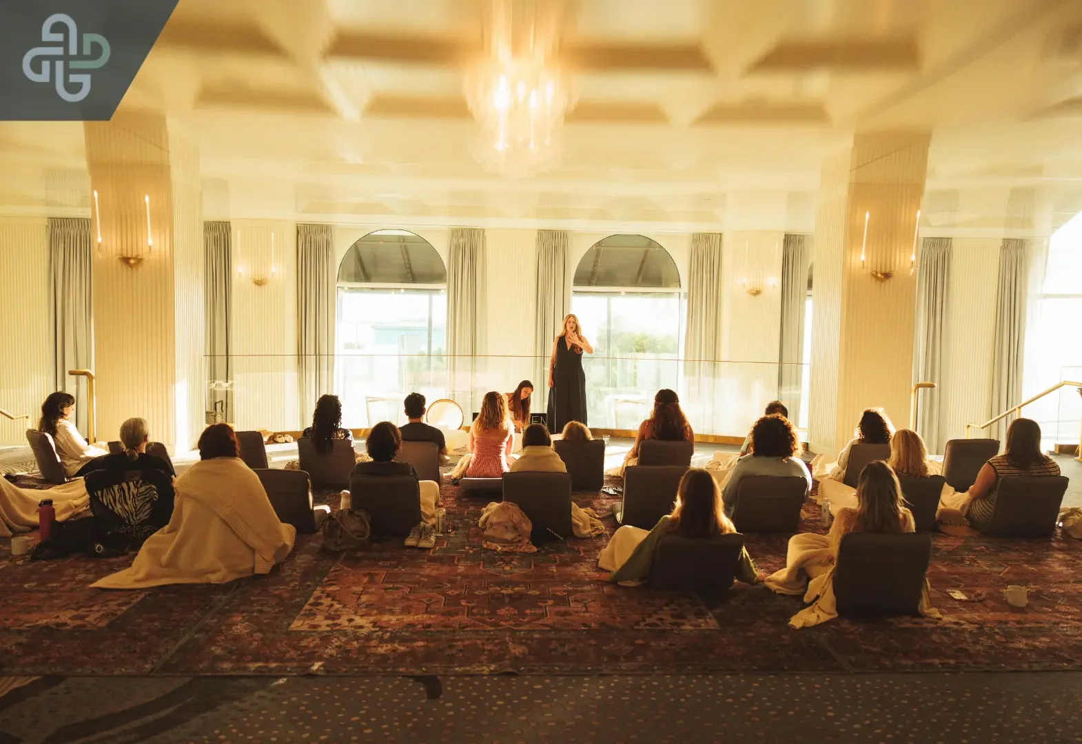 Speaker addresses a seated audience in an elegant, sunlit ballroom with tall windows, chandeliers, and warm golden lighting.