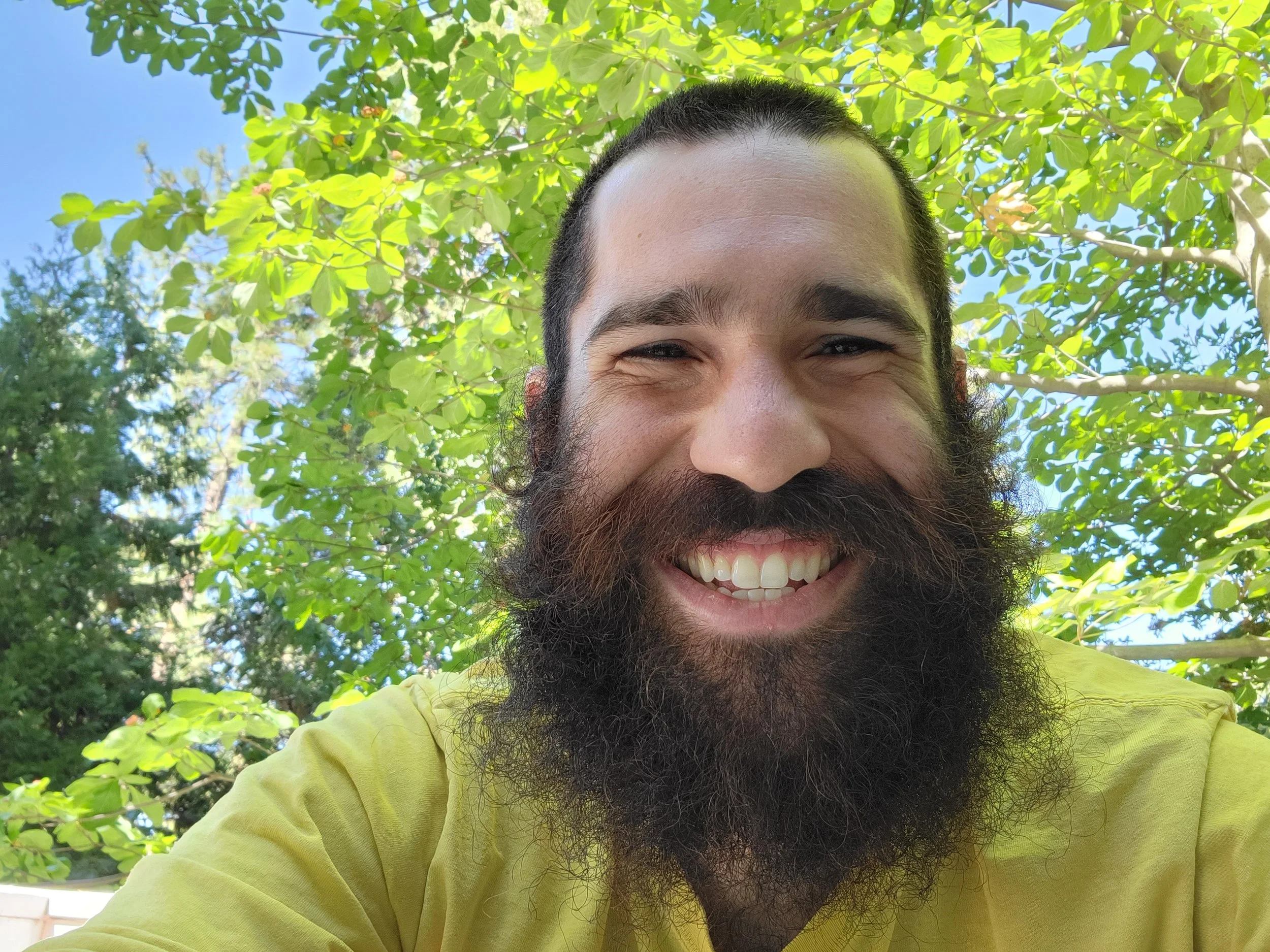Close-up of a smiling man with a beard and dark hair, wearing a yellow shirt, outdoors with green leaves and trees in the background under a clear blue sky.