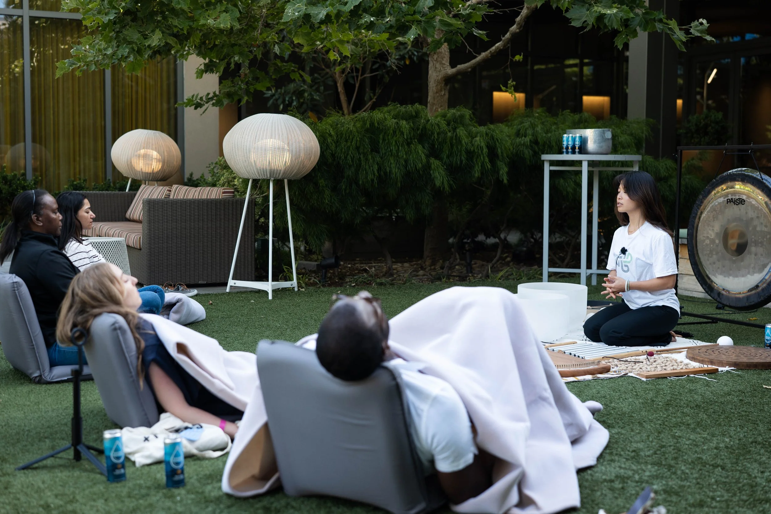 A woman is sitting on the ground in front of an audience, with a gong and singing bowls nearby, leading a meditation or sound therapy session outdoors on a grassy area with trees and modern furniture in the background.