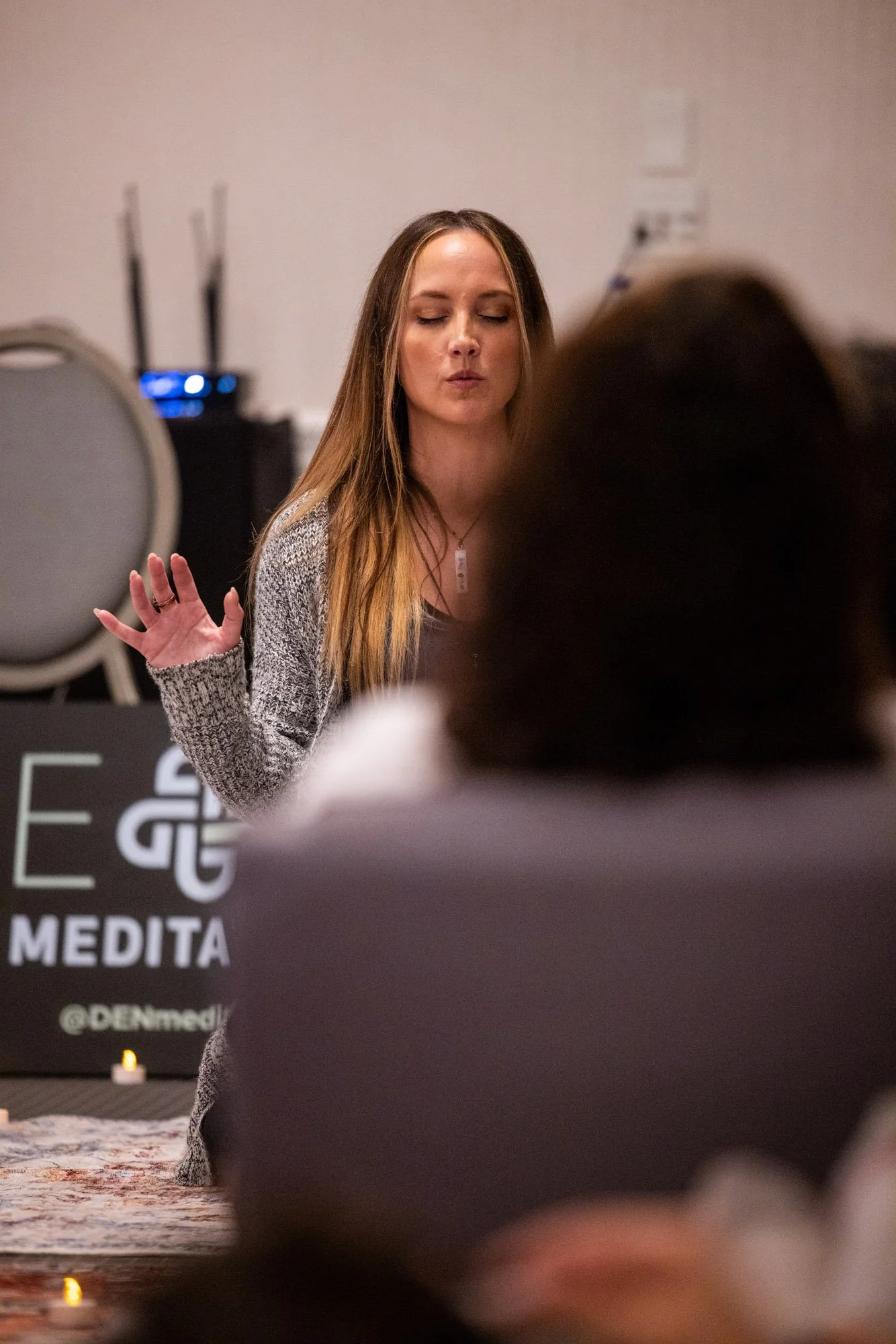 A woman with long brown hair and closed eyes is meditating with her hand raised. She is in a room with stacked chairs and a sign that reads 'MEDITA' in the background.