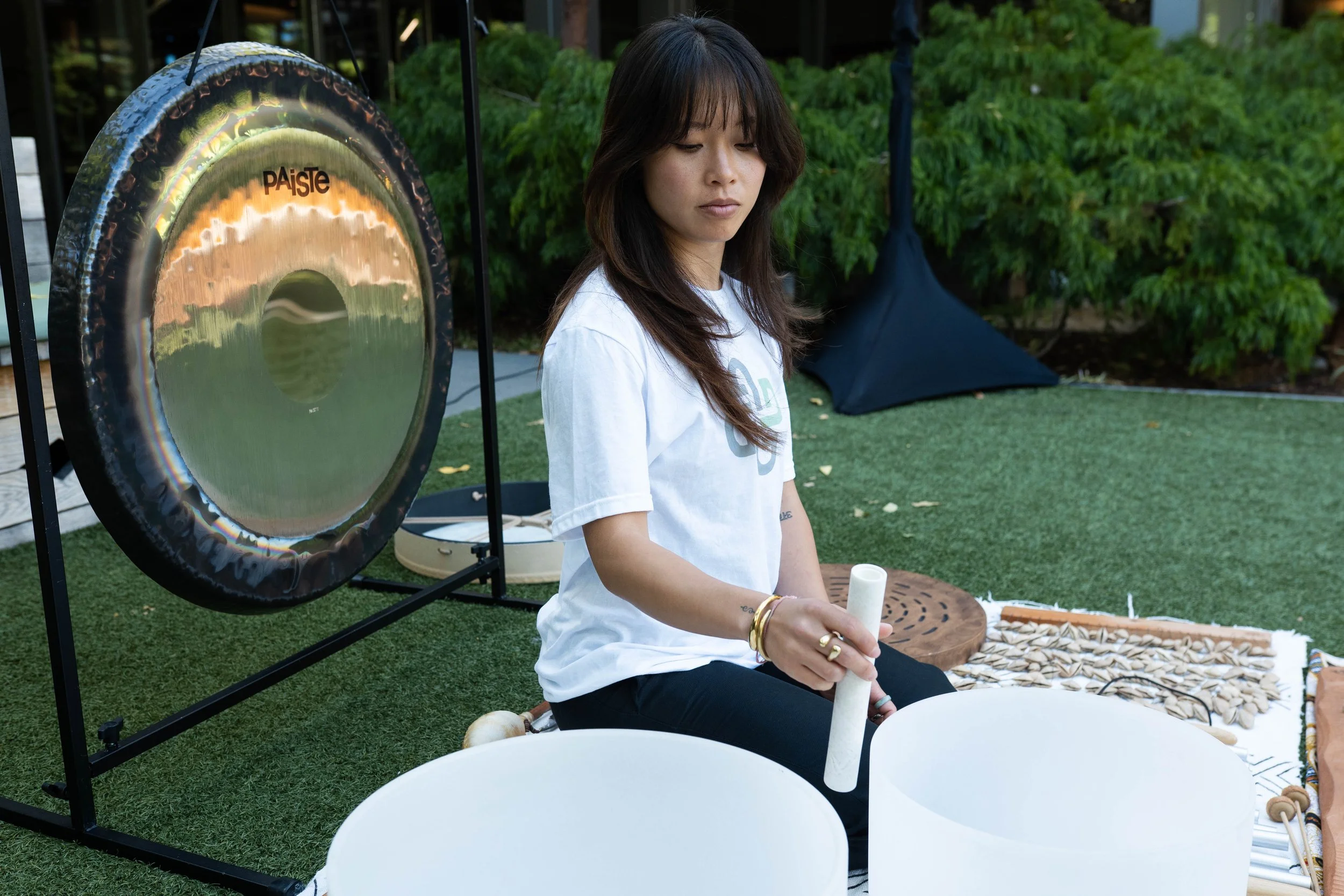 Woman sitting outdoors playing crystal singing bowls with a large gong beside her.
