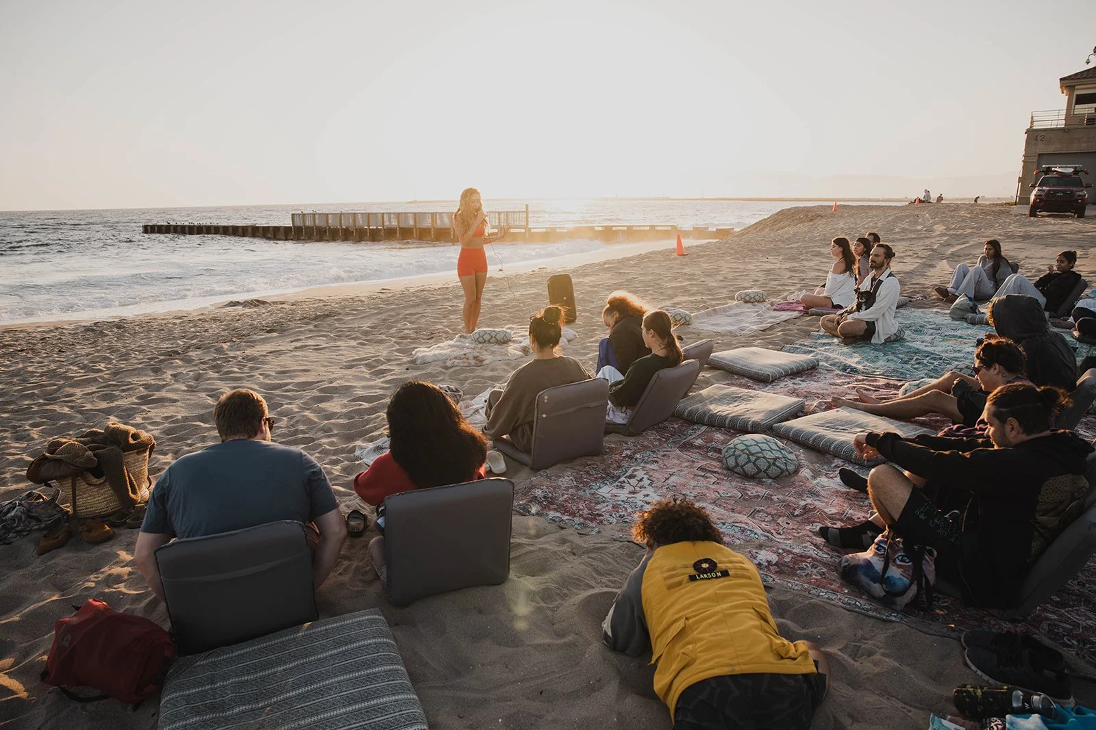 A group of people sitting on cushions, blankets, and chairs on a sandy beach, listening to a woman speaking at a microphone during sunset. The ocean and a pier are visible in the background.