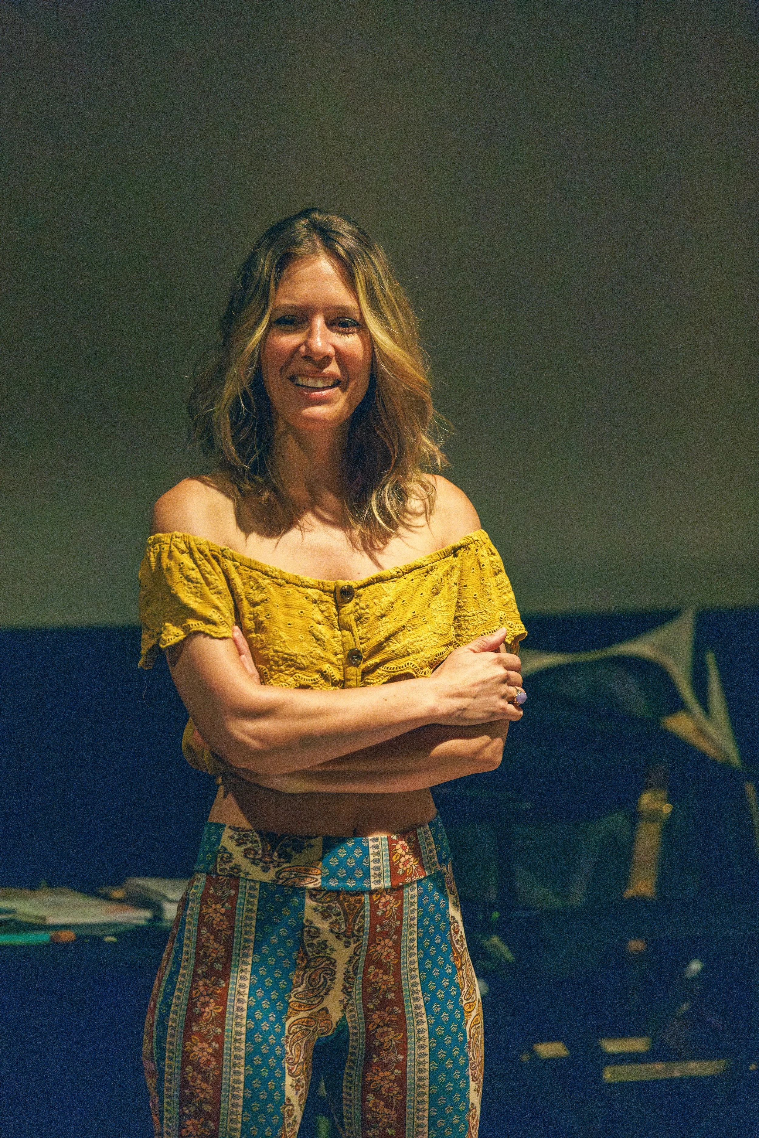 A woman with shoulder-length wavy blonde hair smiling, wearing a yellow off-the-shoulder top and patterned pants, standing in a room with a dark wall background.