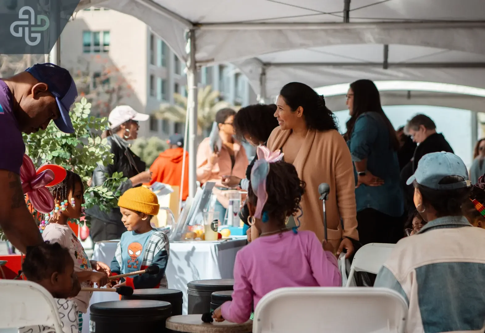Families gather beneath a white canopy at a community event, sharing a table as a smiling woman talks with a child; the scene feels lively and welcoming.