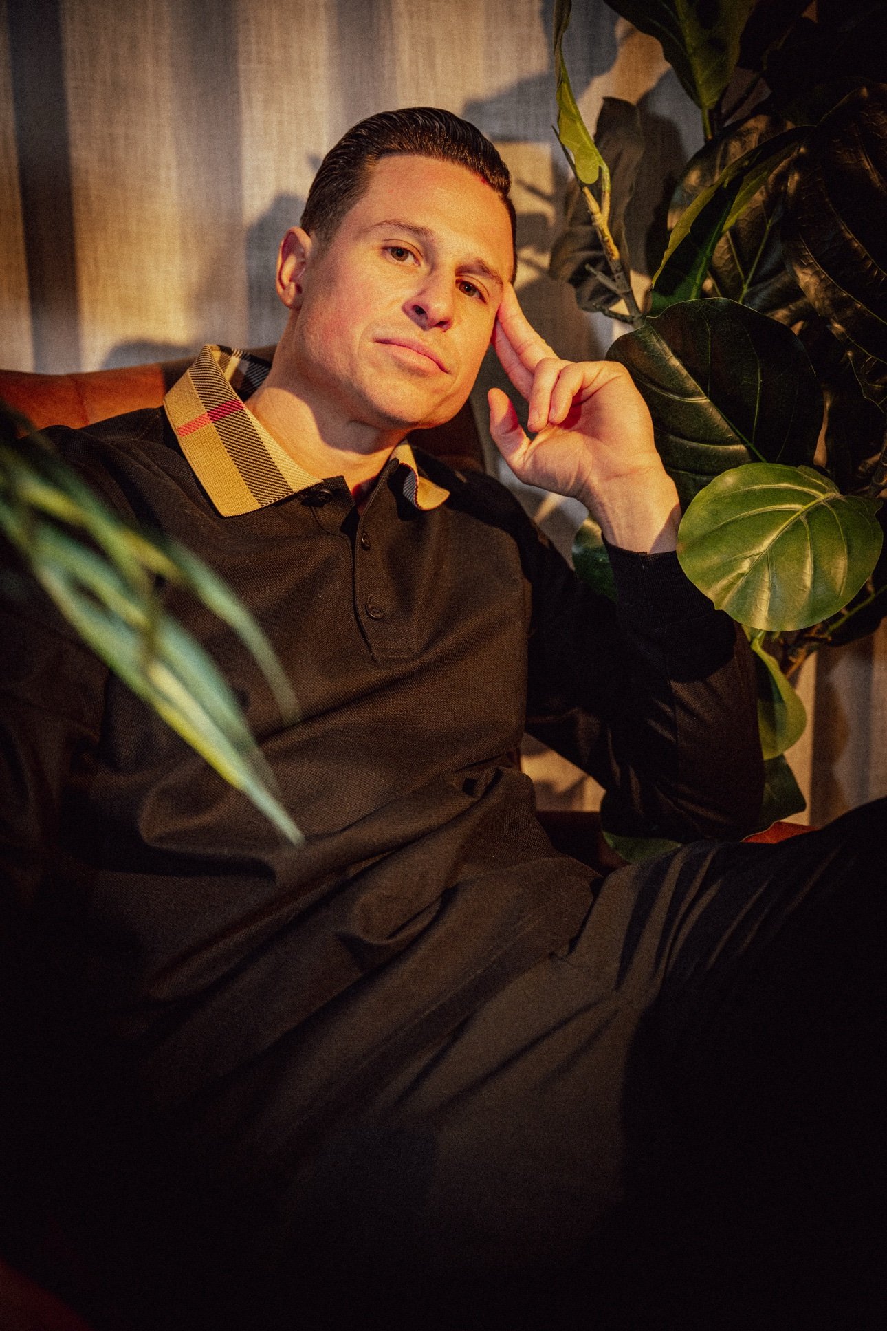 Portrait of a man sitting near green leafy plants with a thoughtful expression, wearing a dark sweater with a checkered collar, in a dimly lit indoor setting.